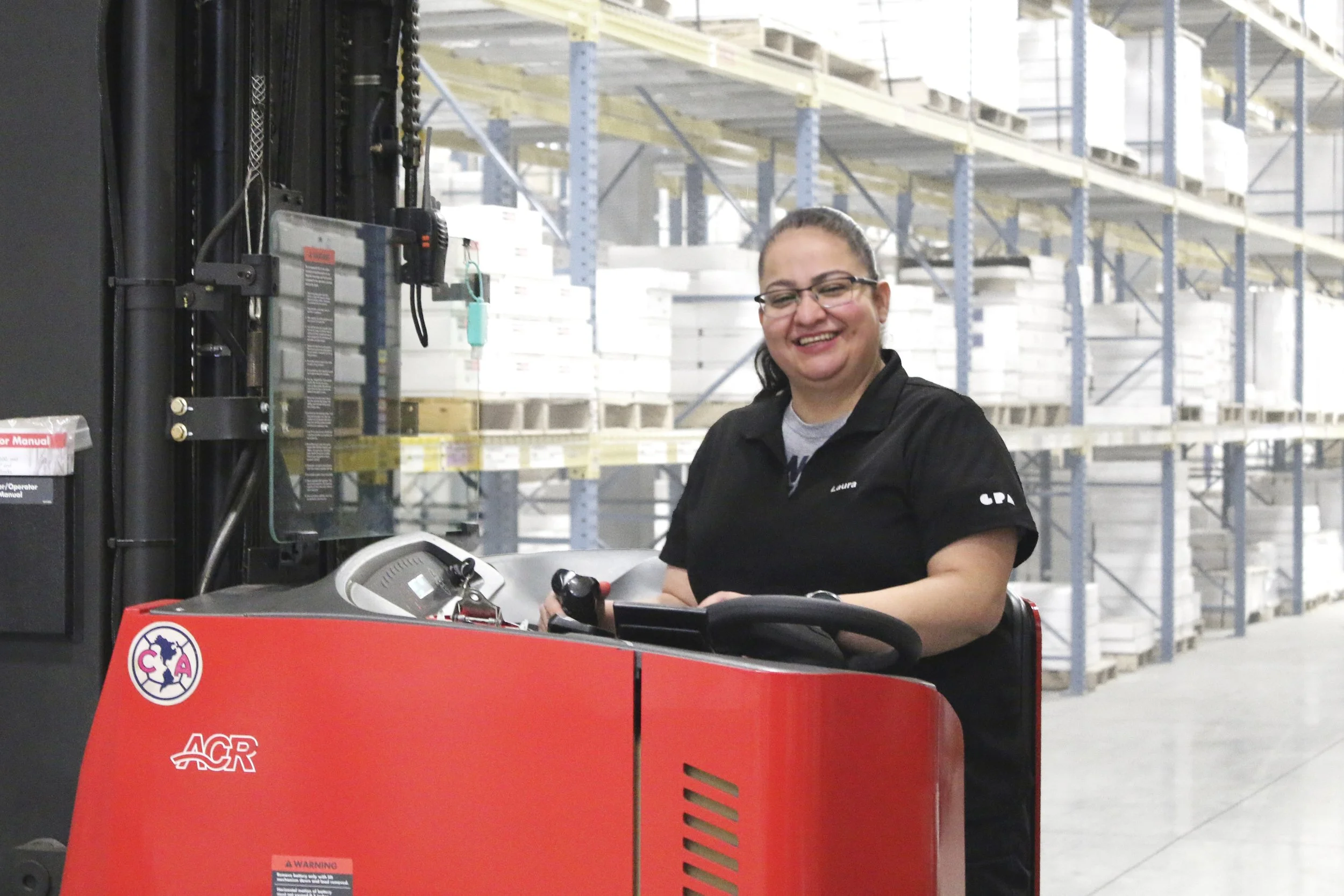 A woman with glasses and a dark uniform smiling at a forklift in a warehouse with metal shelving units filled with boxes in the background.