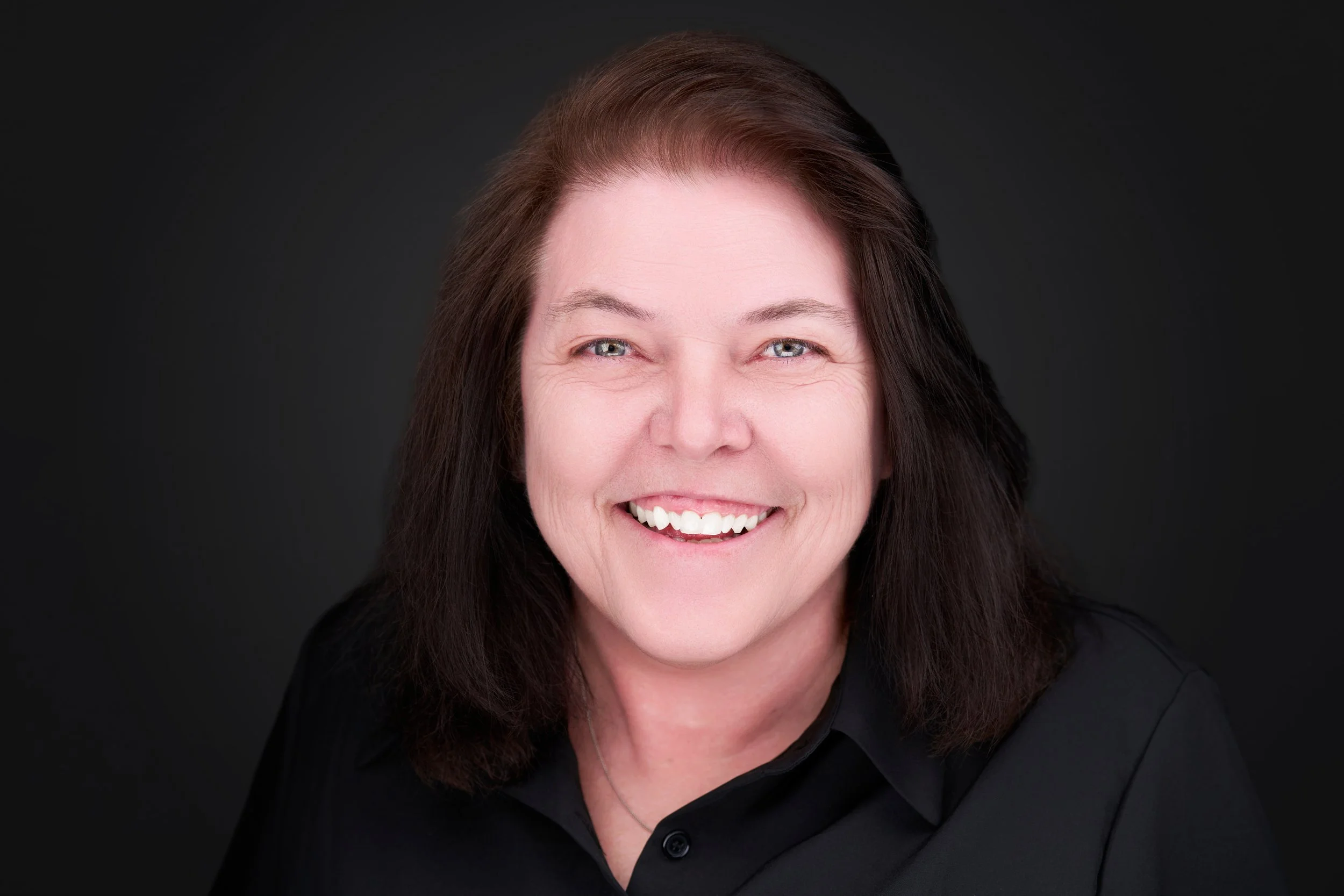 A smiling middle-aged woman with long dark brown hair and blue eyes, wearing a black top, against a dark background.
