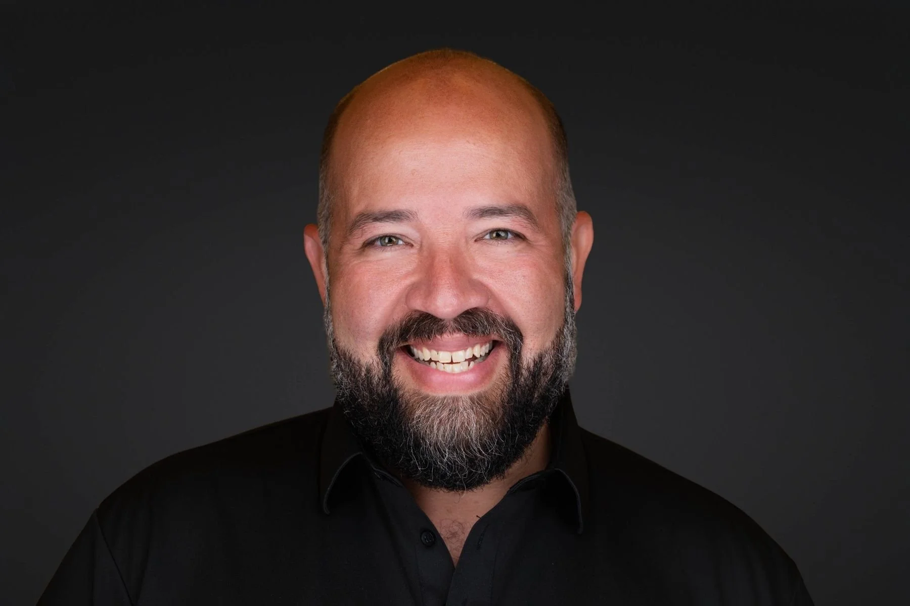A smiling man with a bald head, a full beard, wearing a black shirt and standing against a dark background.