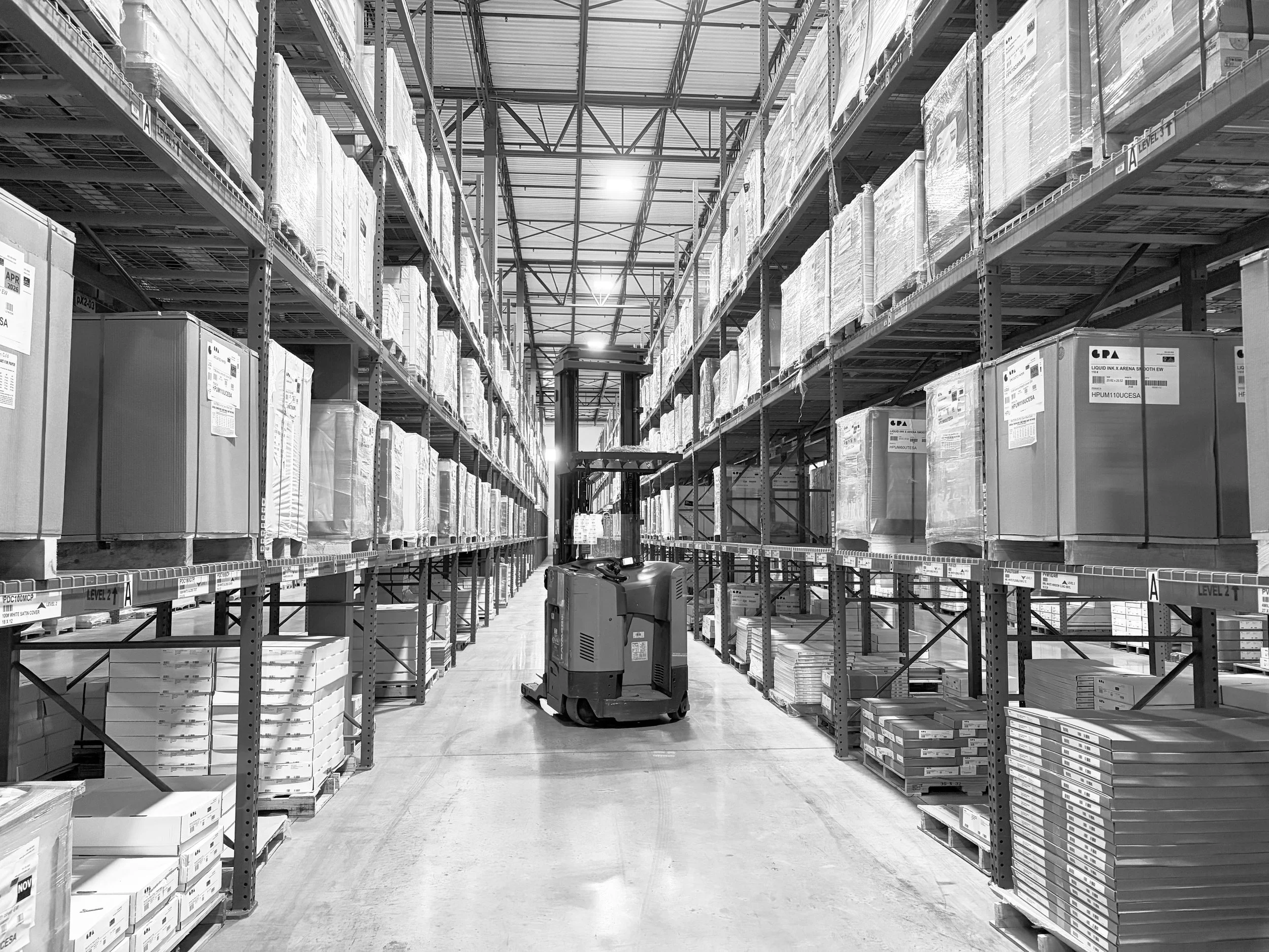 Warehouse aisle with shelves stacked with boxes, a forklift in the center, and bright lighting from the ceiling.