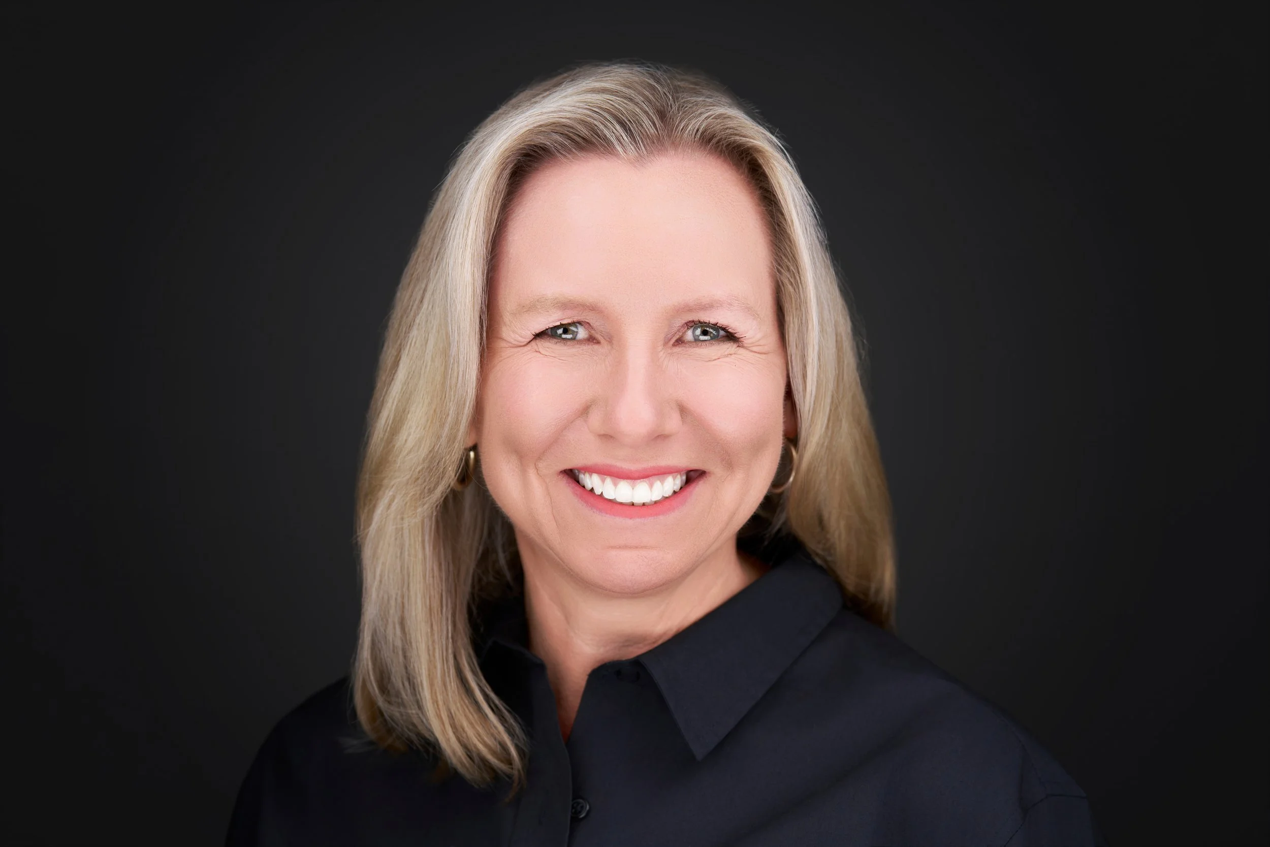 Close-up portrait of a smiling middle-aged woman with long blonde hair, wearing a dark top, against a dark background.