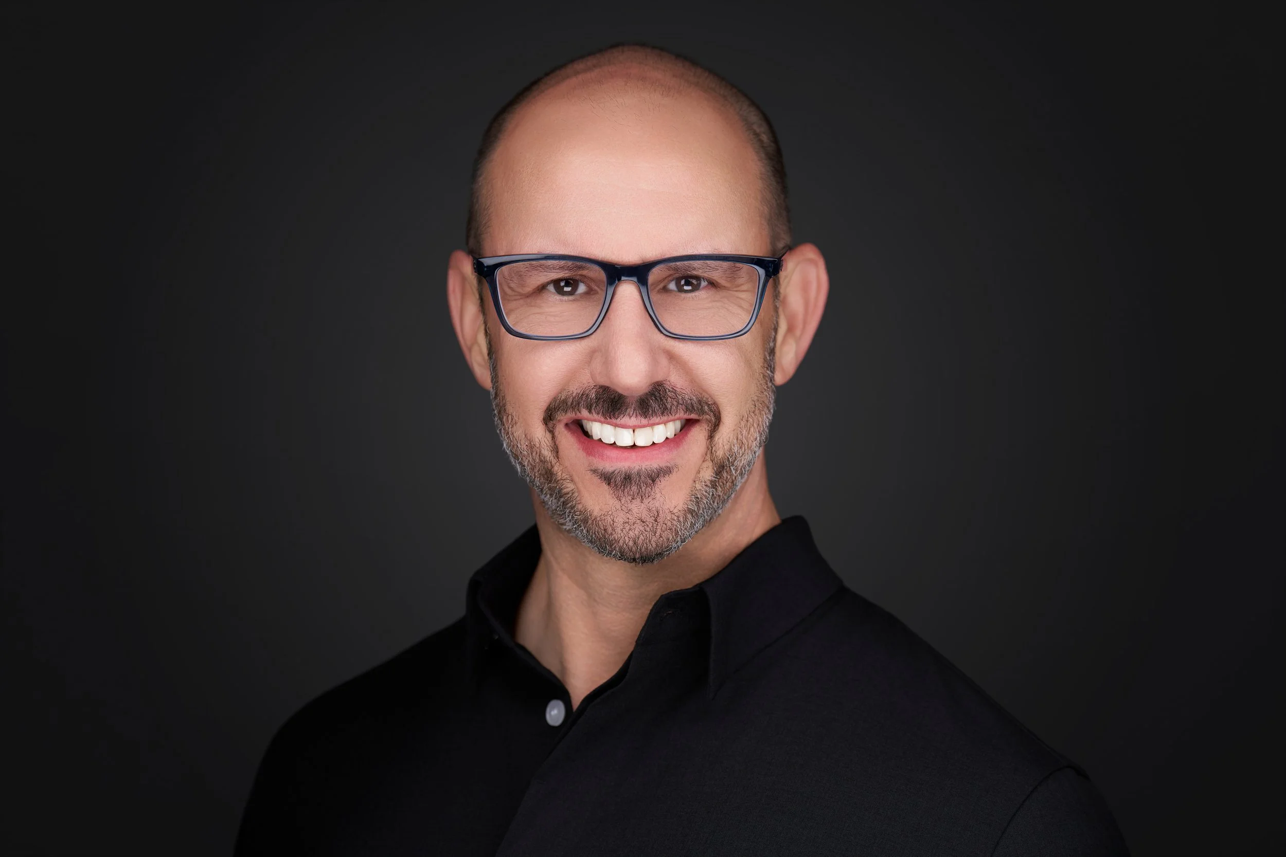A man with a shaved head and glasses smiling, wearing a black shirt, against a dark background.