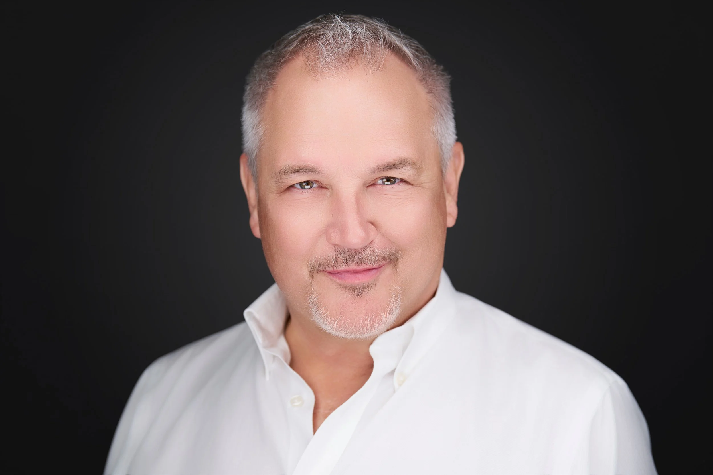 Close-up portrait of a middle-aged man with short gray hair, light facial hair, and a slight smile, wearing a white collared shirt against a dark background.