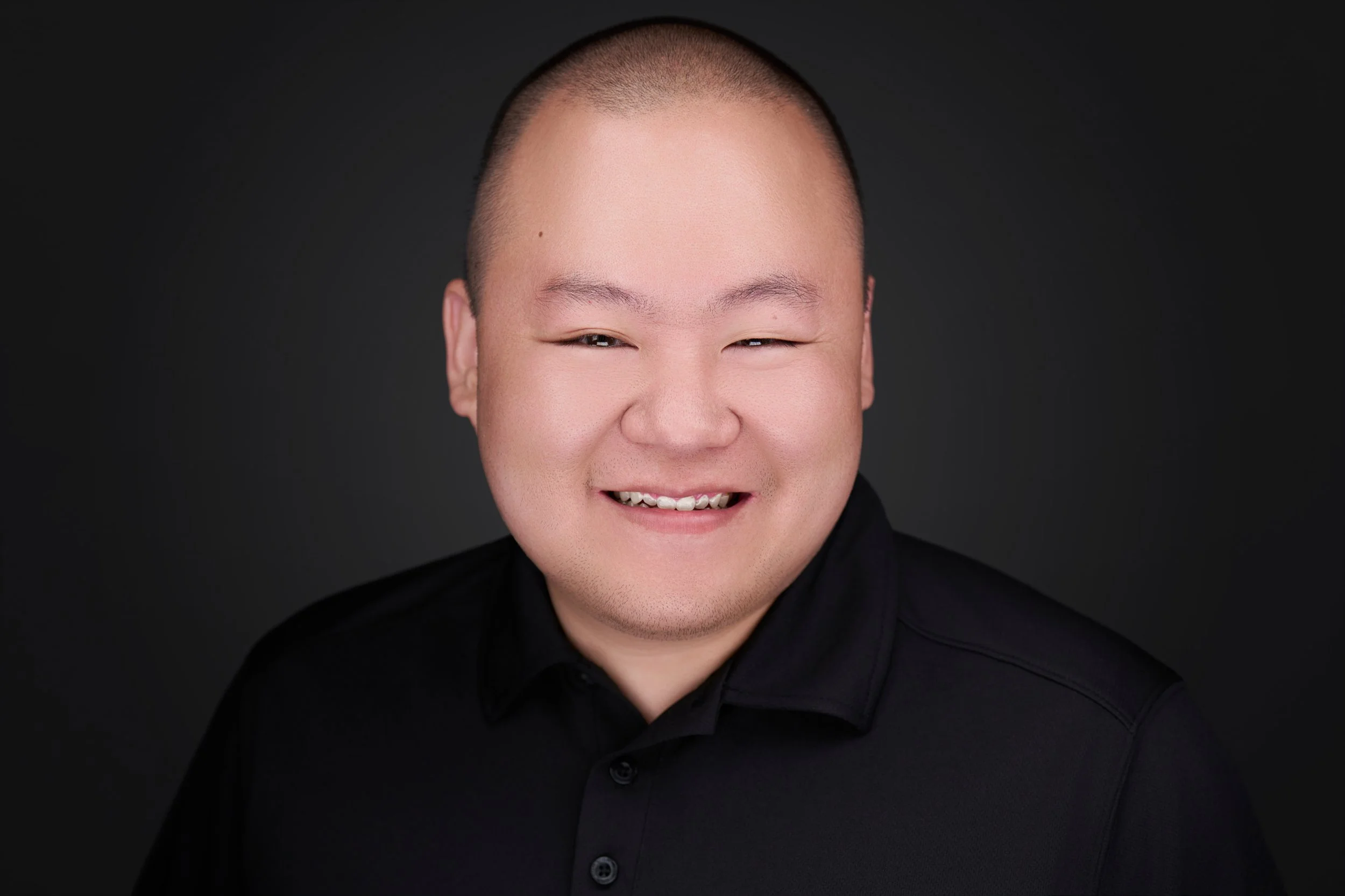 Close-up of a smiling man with a shaved head wearing a black shirt against a dark background.