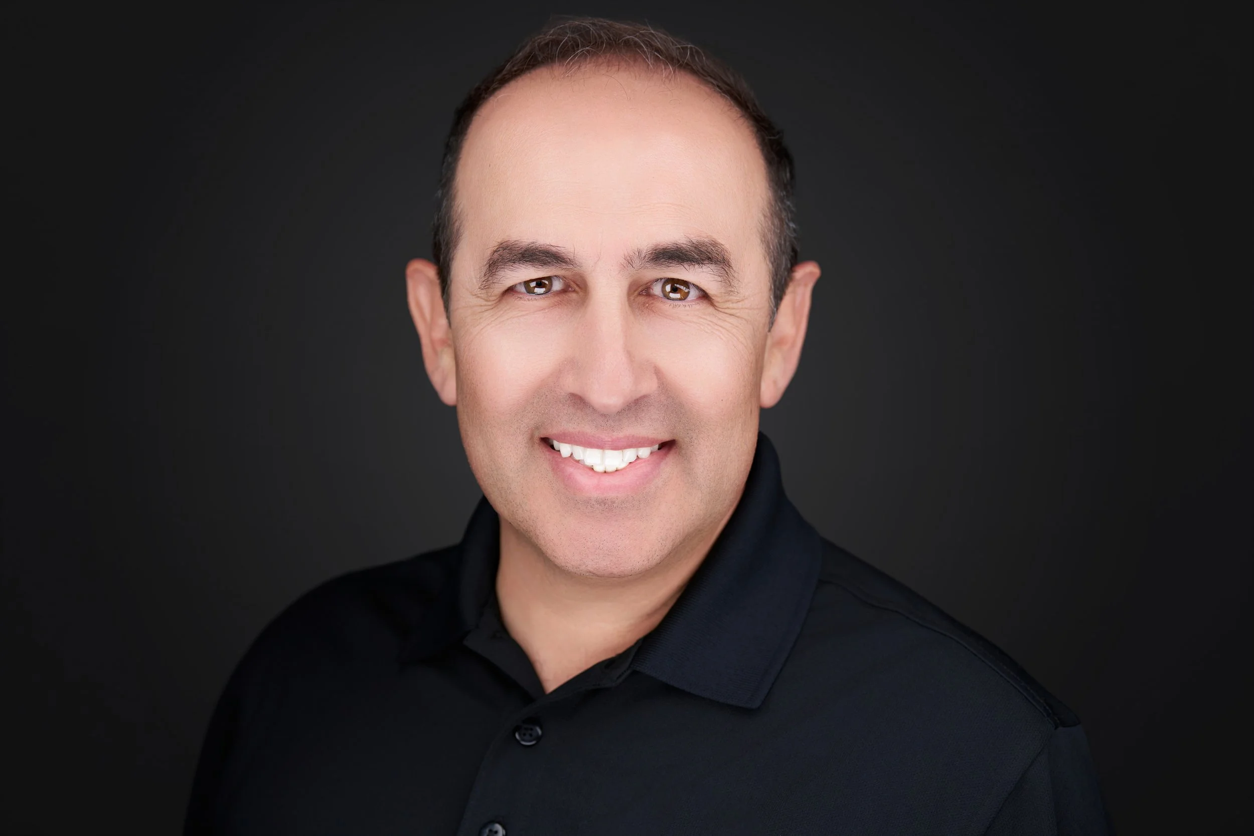 Headshot of a smiling man with short brown hair, brown eyes, and a light complexion, wearing a black shirt against a dark background.
