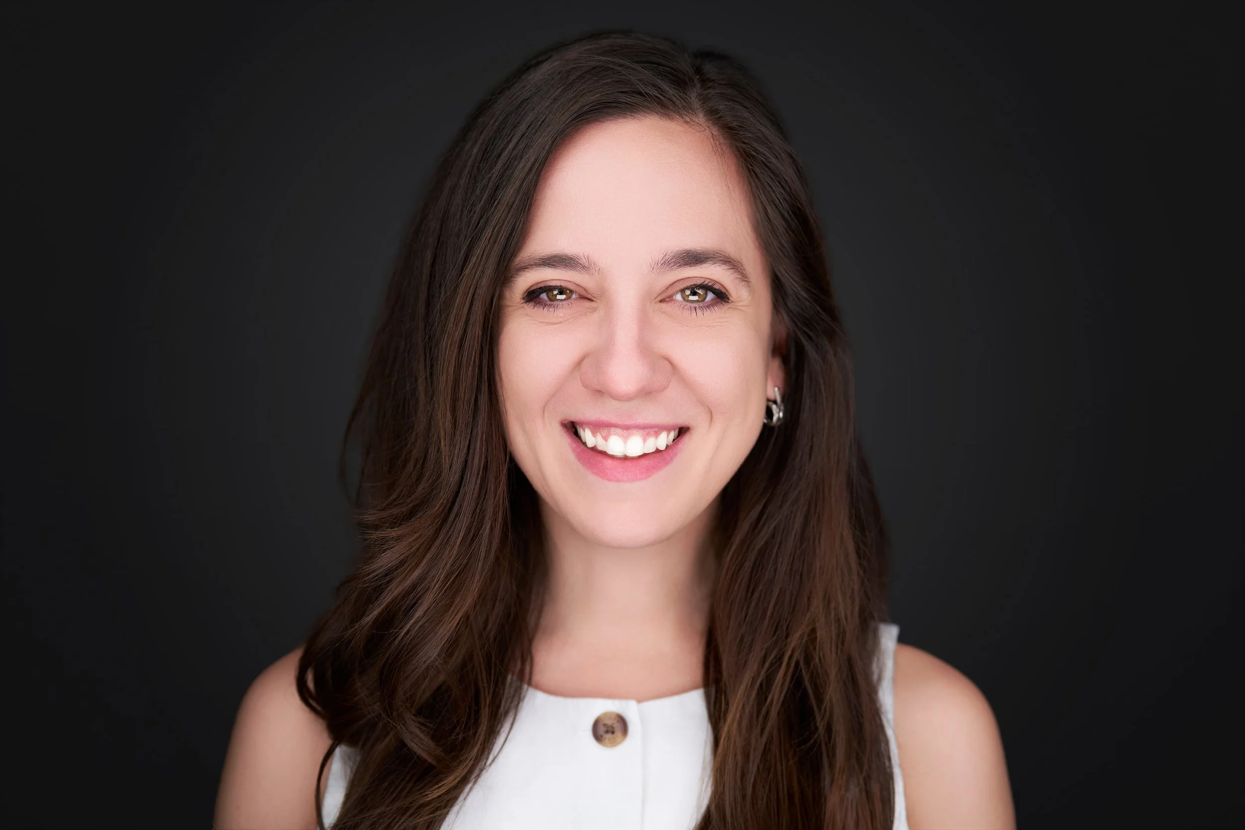 A woman with long brown hair, smiling, wearing a white sleeveless top with a button, and earrings, against a black background.