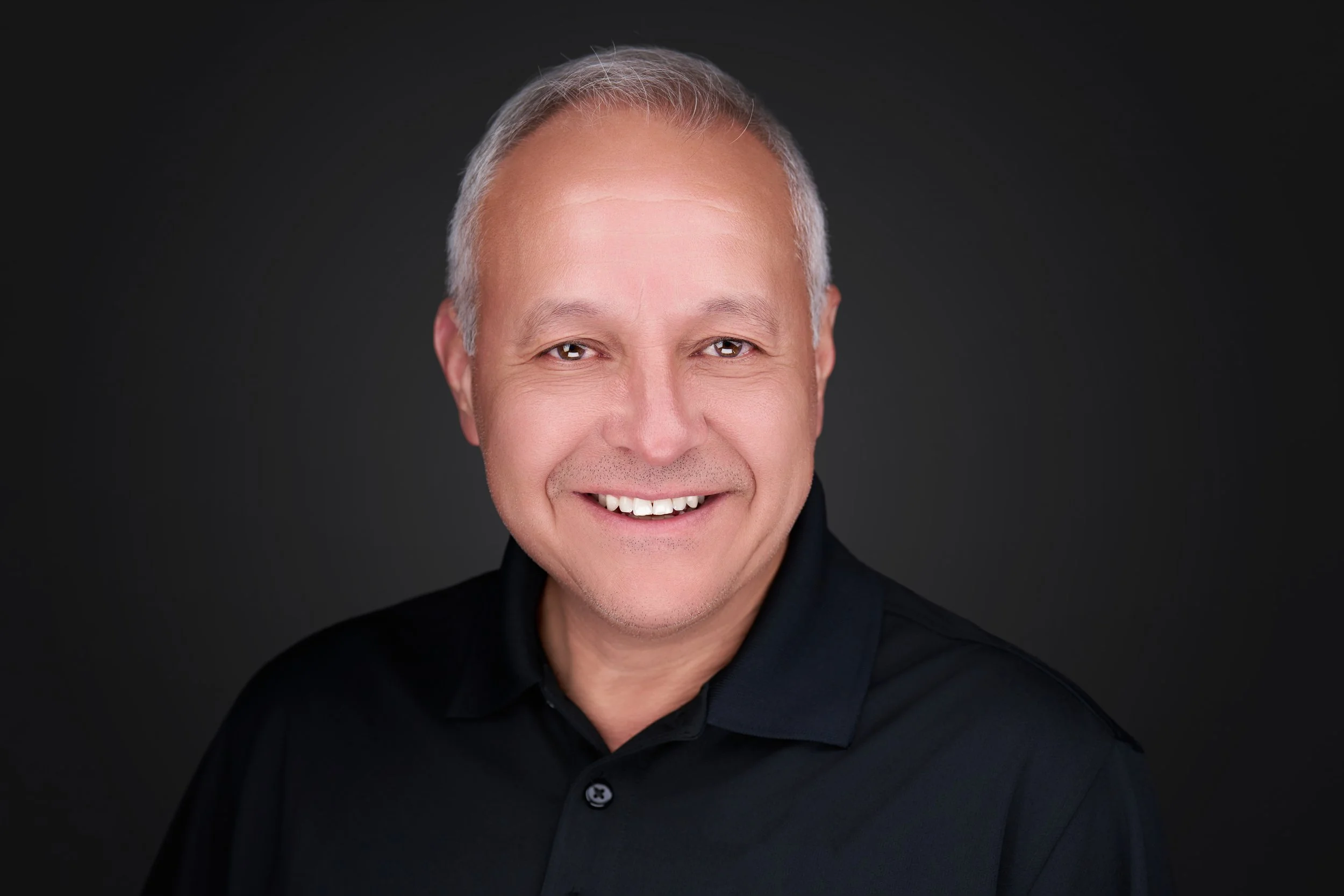 Close-up portrait of a middle-aged man with gray hair, smiling, wearing a black shirt against a dark background.
