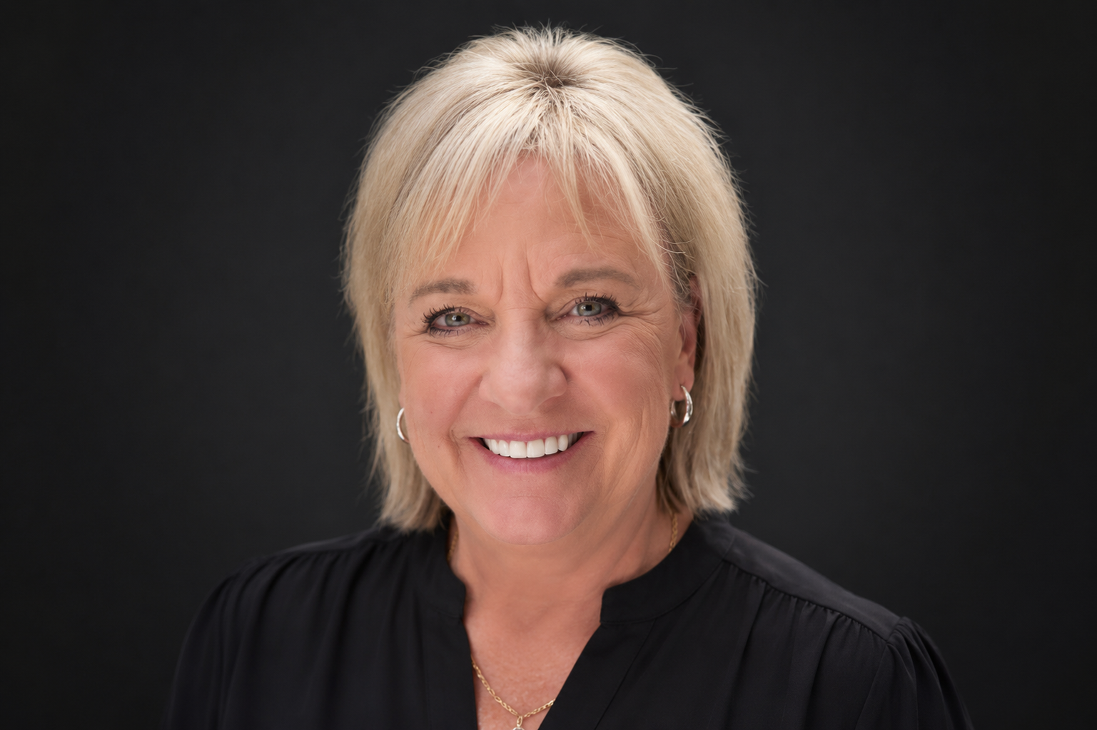 A portrait of a smiling middle-aged woman with short blonde hair, hoop earrings, wearing a black top, against a black background.