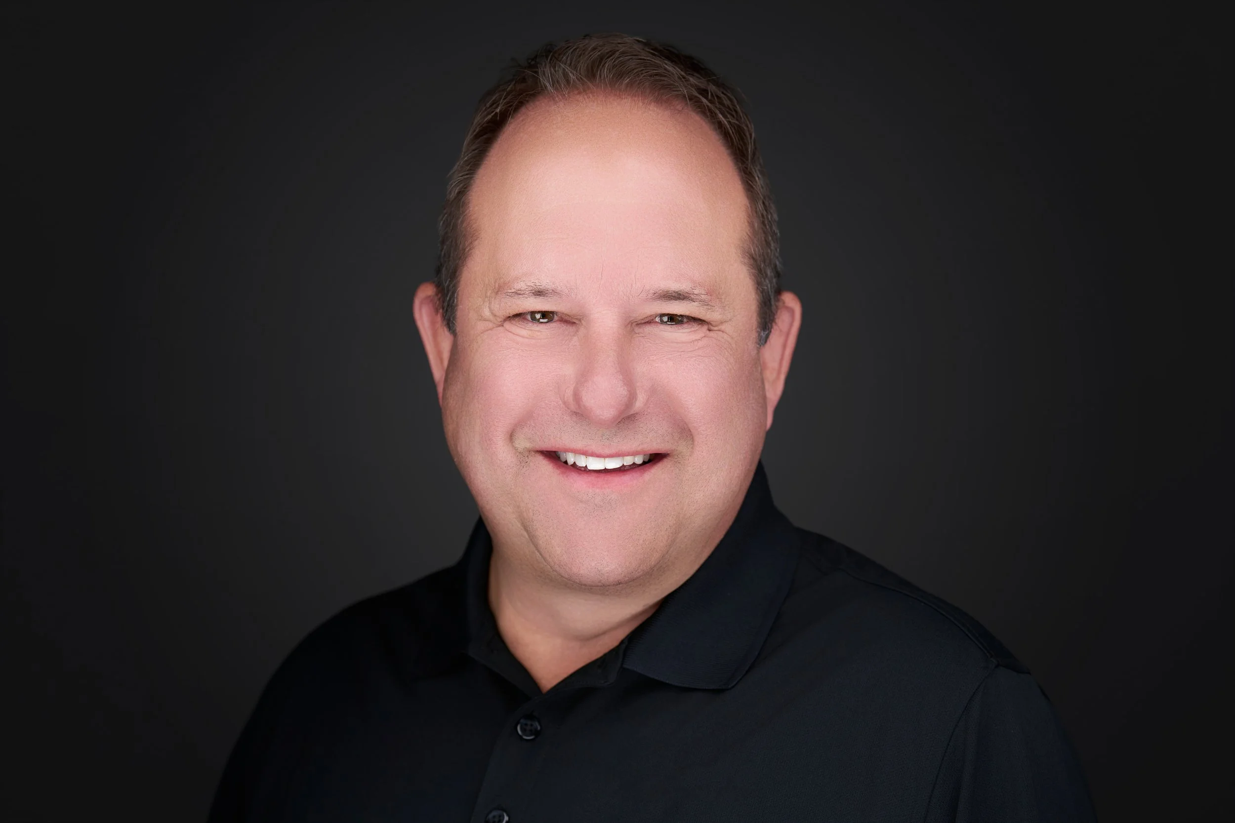 A smiling man with short brown hair, wearing a black shirt, against a dark background. Barry Nelson