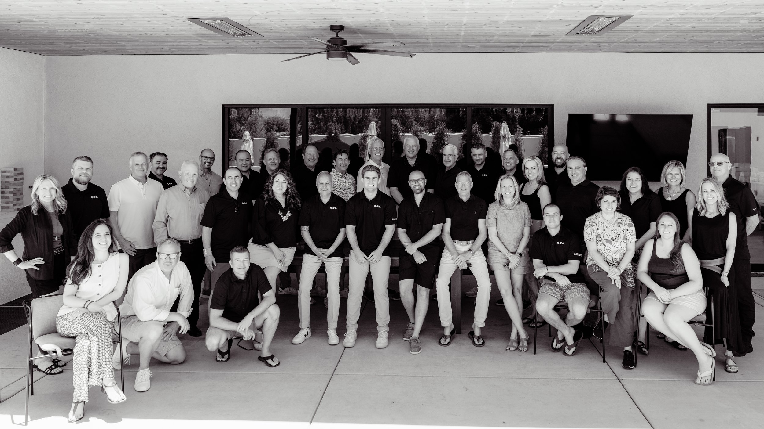Group photo of 30 people, some in black shirts, indoors with a large window and a TV screen behind them.