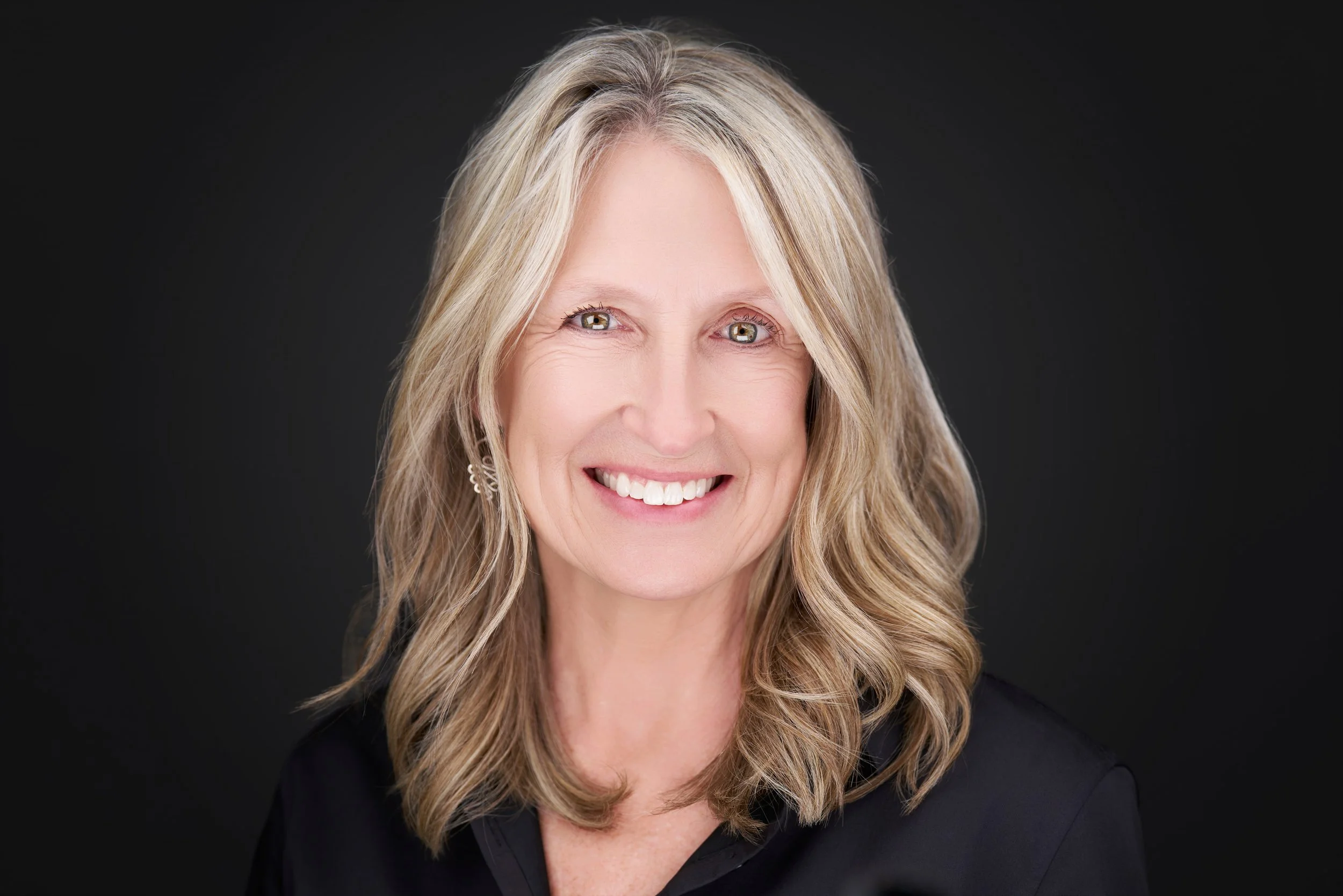 Headshot of a middle-aged woman with long wavy blonde hair, smiling, wearing a black top, against a dark background.