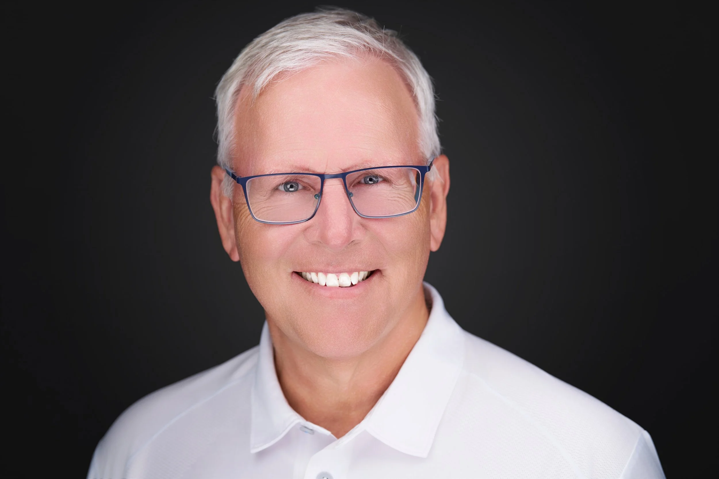 A smiling man with gray hair, wearing glasses and a white shirt, against a dark background.