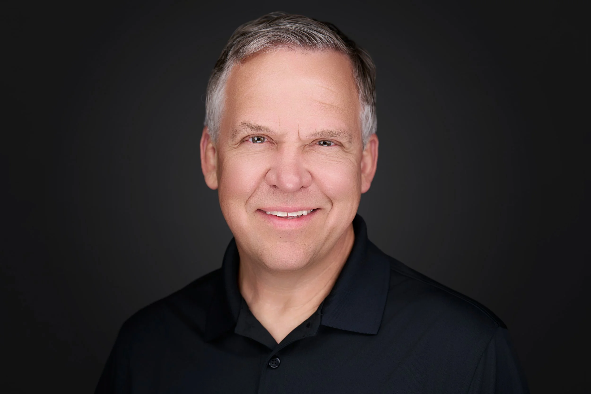 A middle-aged man with short, gray hair wearing a black shirt, smiling against a dark background.