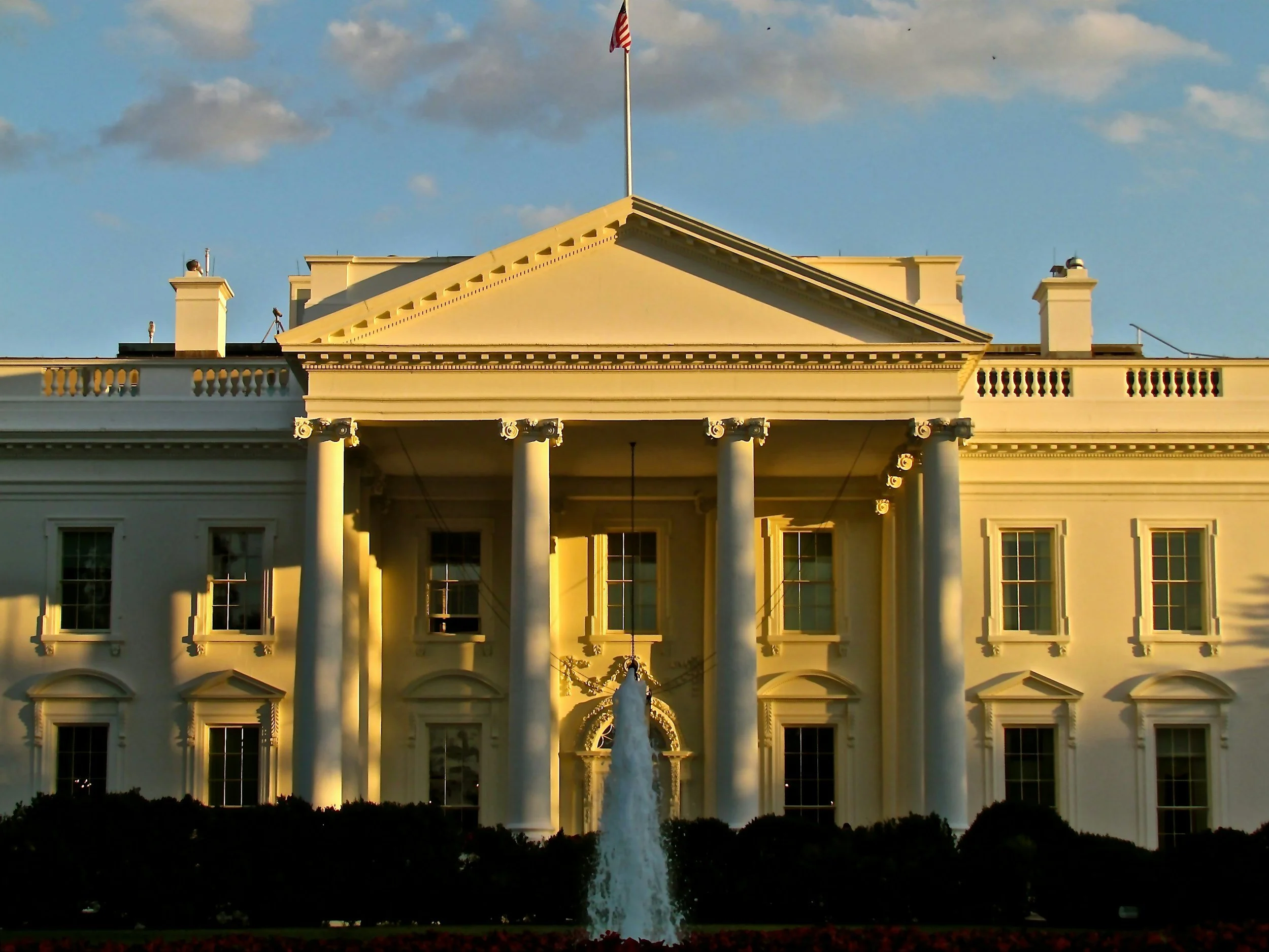 The White House bathed in golden sunset light, with a fountain in the foreground and the American flag on the roof, representing Schedule C and Schedule G hiring authorities, which are used for political appointments to the federal government.