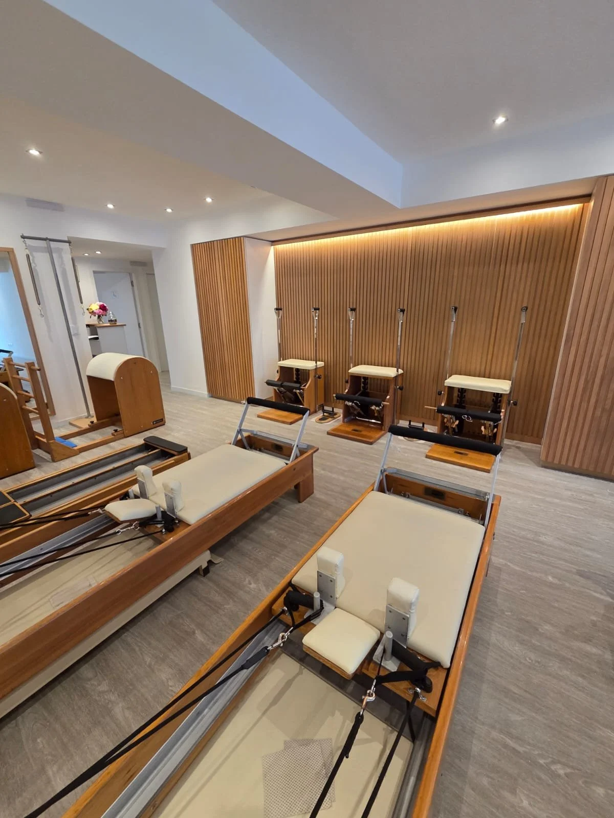Two Pilates reformer machines and four vertical Pilates chairs in a modern, wooden-floored Pilates studio with wood-paneled wall and recessed ceiling lights.