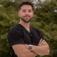 A man with short dark hair and a beard, wearing a black shirt, standing outdoors with arms crossed and smiling, with green trees in the background.