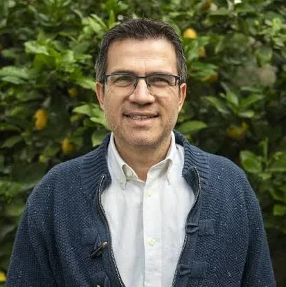 A man with dark hair, glasses, and a light shirt standing outdoors in front of a green leafy background.