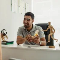 A man sitting at a desk with a smile, holding a model of a human brain, surrounded by artistic sculptures and a green notebook.