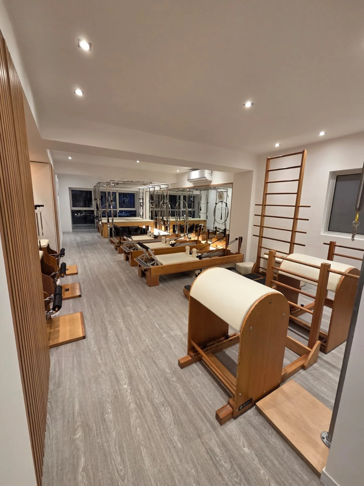 Pilates studio with wooden equipment, mats, and mirrors, illuminated by ceiling lights.
