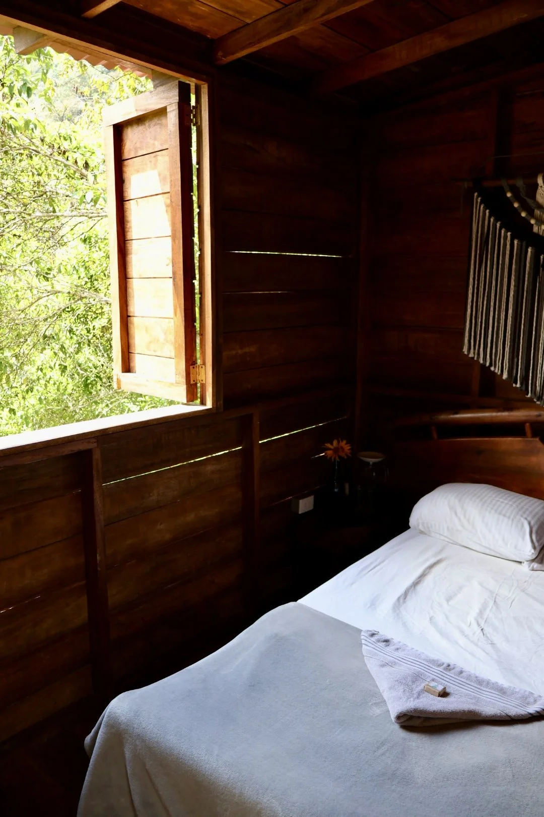 A wooden bedroom with an open window showing green foliage outside. The bed has white sheets, a pillow, and a folded towel with a small soap bar on top. There is a small bedside table with a flower in a vase.