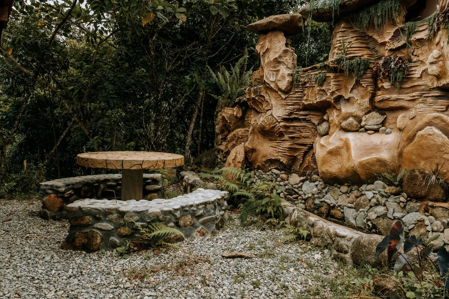 A small outdoor area with a round wooden table and a stone bench surrounded by greenery, including ferns, with a textured rock wall in the background.