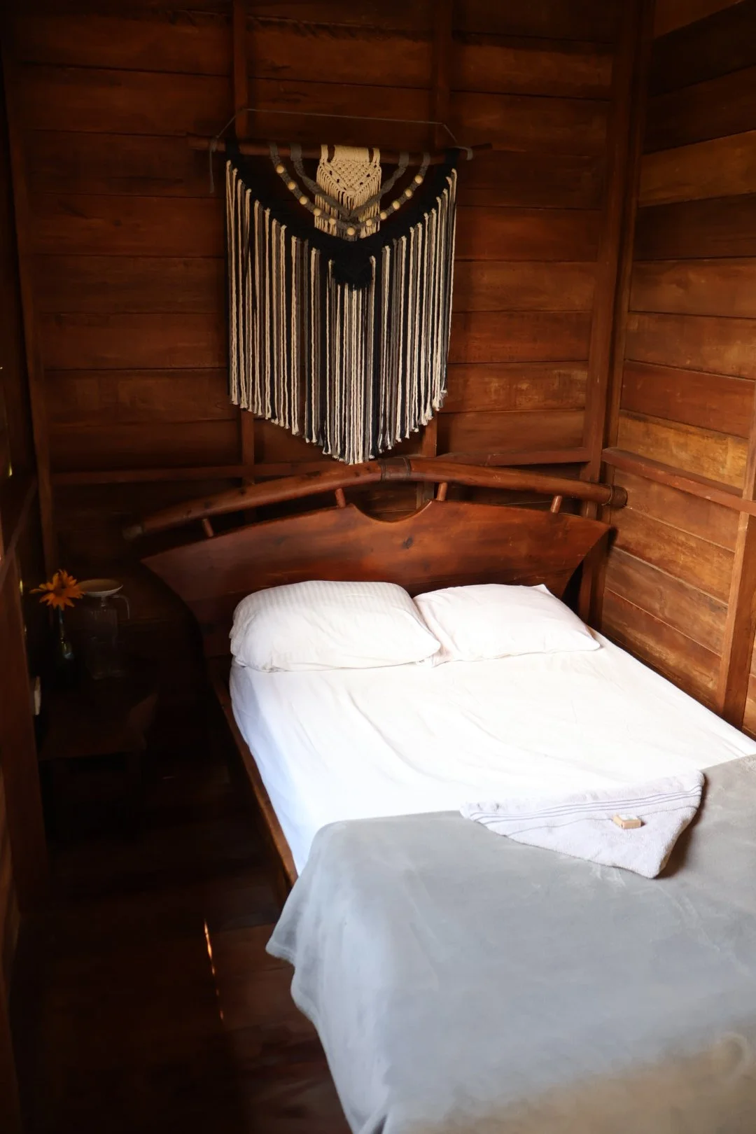 A rustic bedroom with wooden walls, a wooden bed frame, a white sheet, pillows, a nightstand with a small flower vase, and a black and white macramé wall hanging.