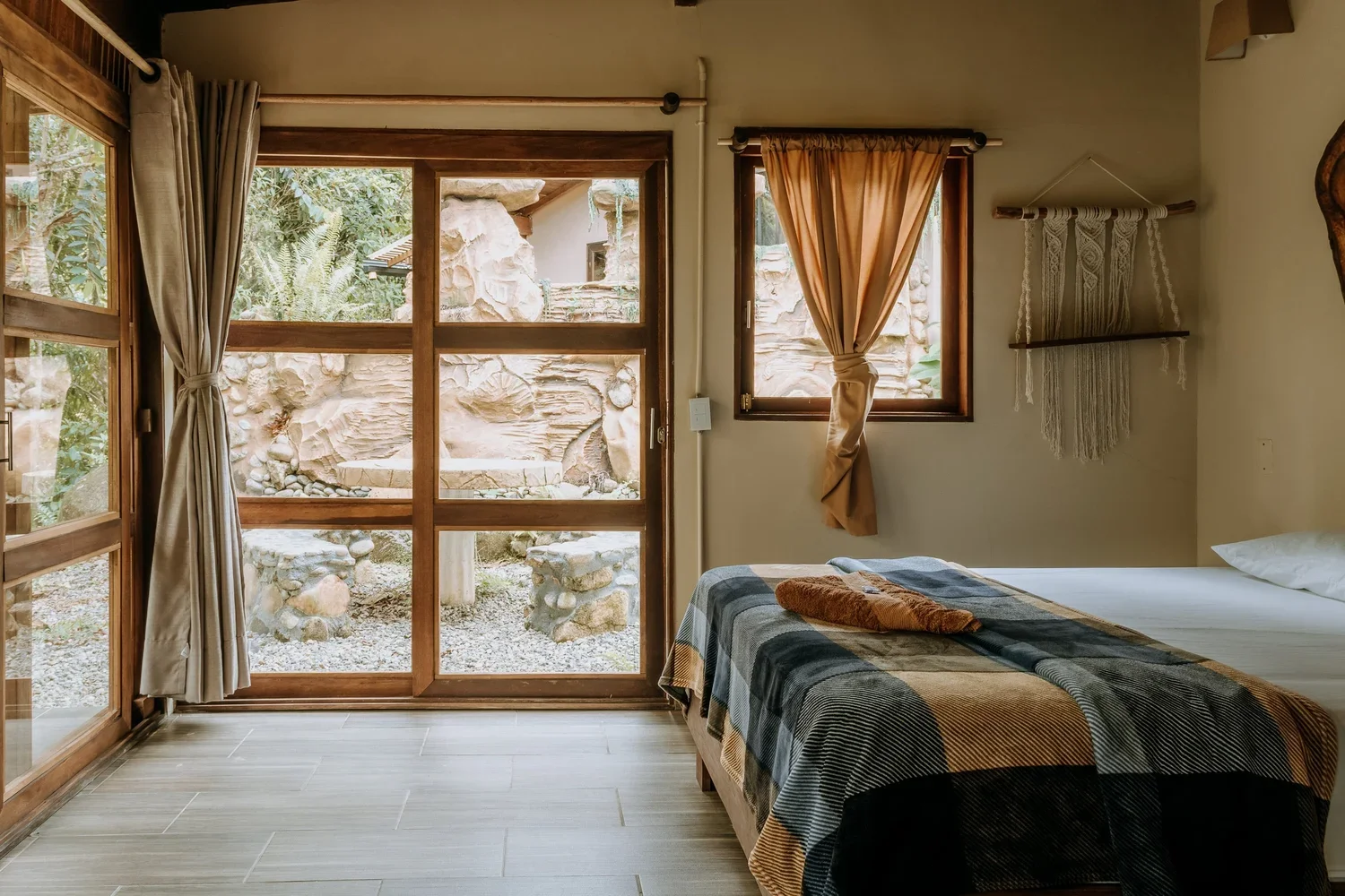 Bedroom with large glass sliding door and window, view of rock garden outside, bed with plaid blanket and towel, and macrame wall hanging.
