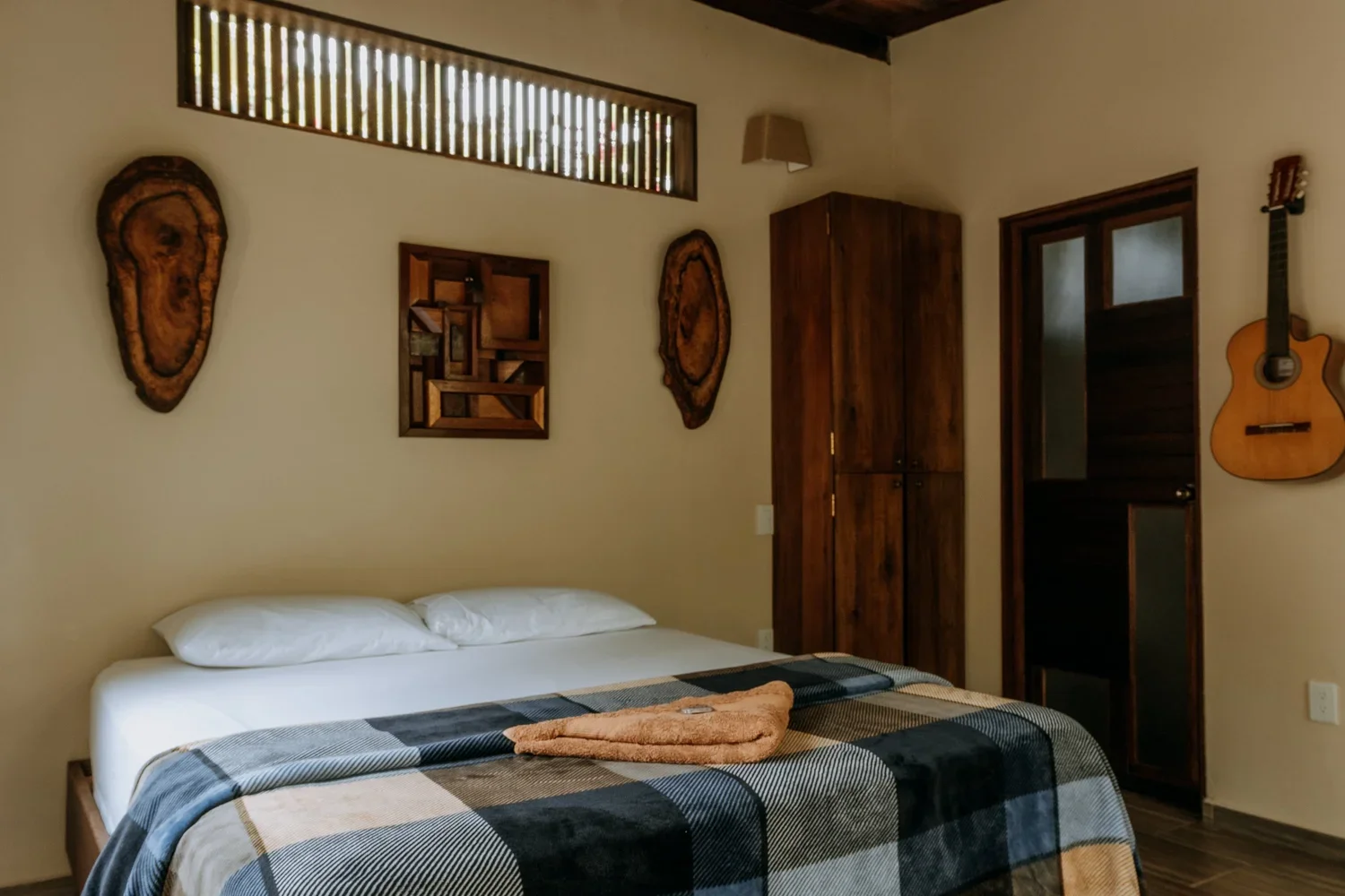 A bedroom with a bed covered by a checkered blanket and pillows, wooden wall decor, a wooden wardrobe, a guitar hanging on the wall, and a window high on the wall.