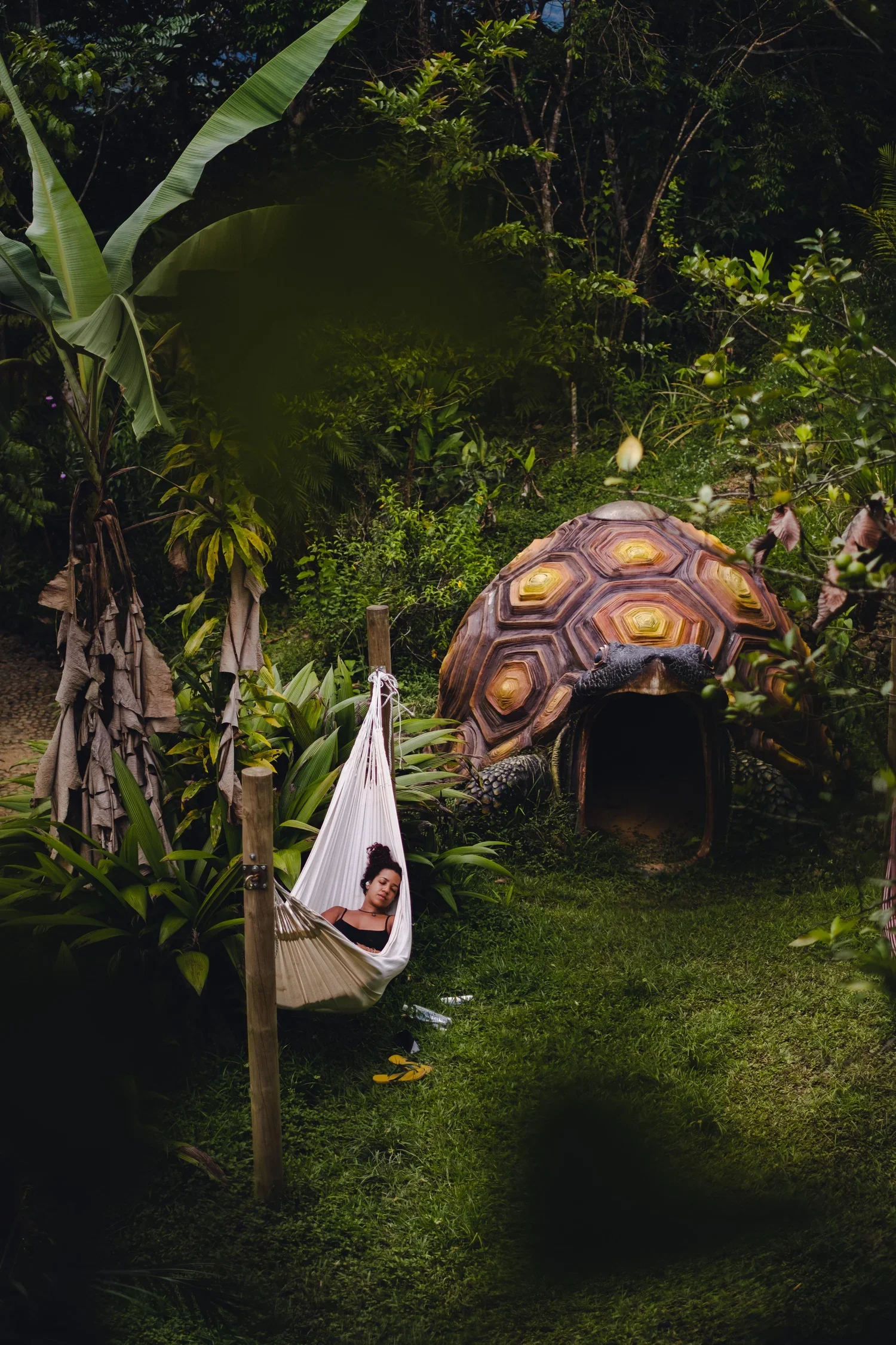 A woman sleeping in a hammock in a lush tropical garden with a small turtle-shaped entrance to a structure in the background.