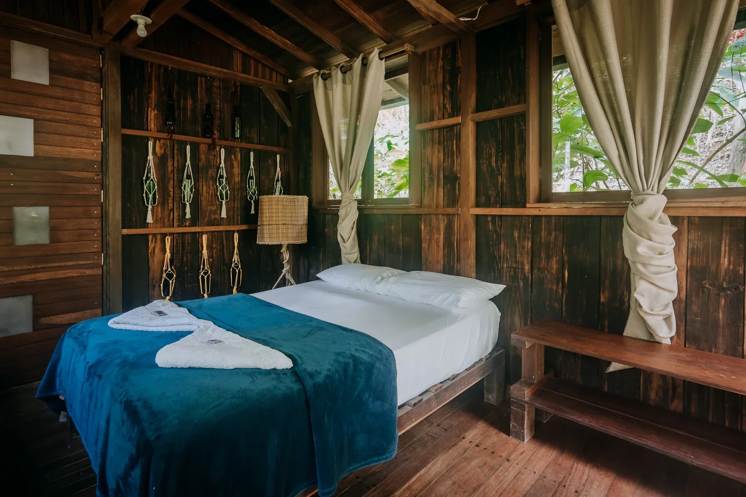 Interior of a rustic wooden cabin bedroom with a bed, white sheets, blue blanket, two towels, a lamp, and windows with beige curtains.