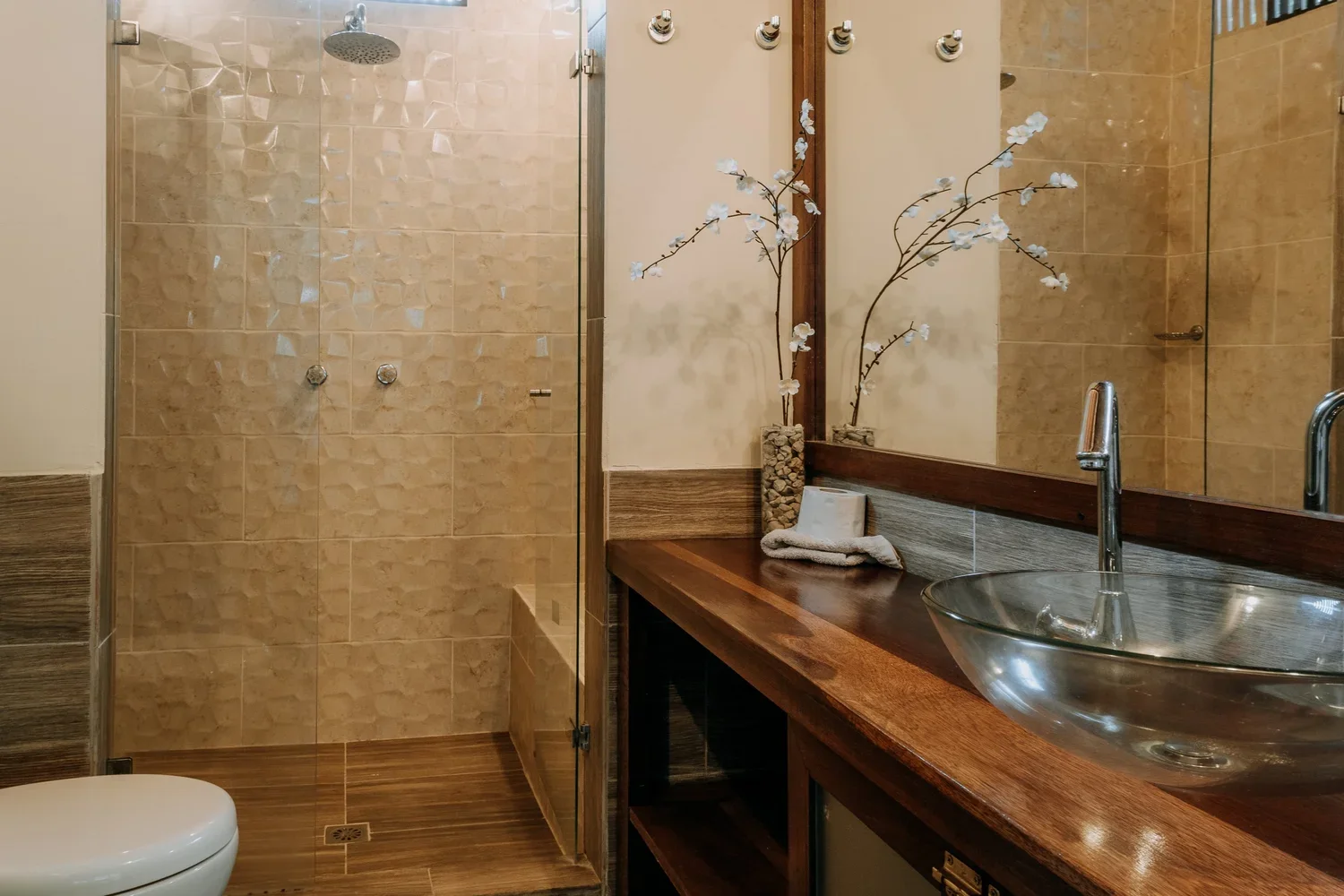 Modern bathroom with a glass shower enclosure, a round glass sink on a wooden countertop, a faucet, a large mirror, decorative flowers, and a toilet.