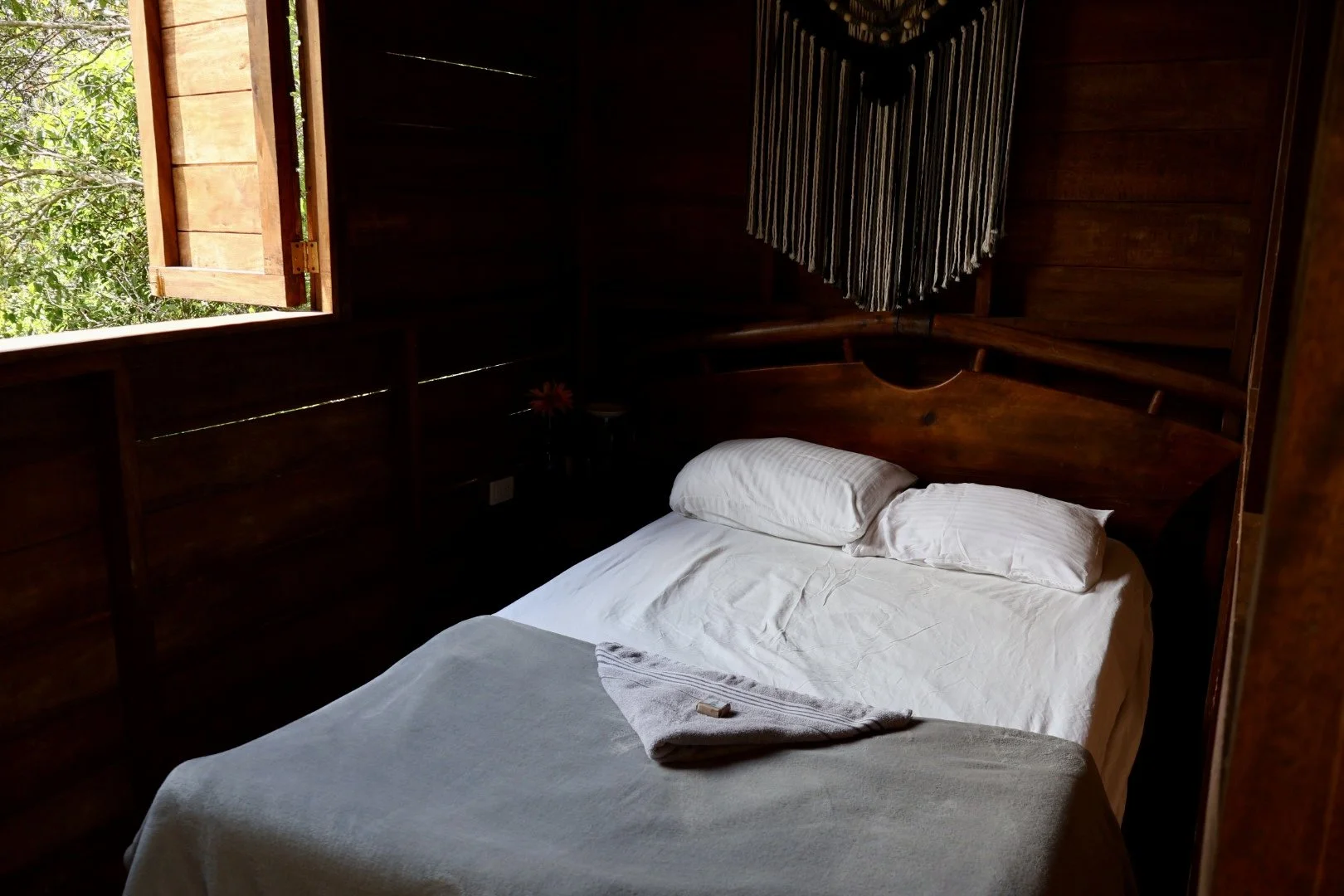 A cozy bedroom with a wooden bed, white sheets, pillow, and a folded blanket. An open window with wooden shutters lets in natural light, revealing green foliage outside. A decorative wall hanging decorates the dark wood-paneled wall.