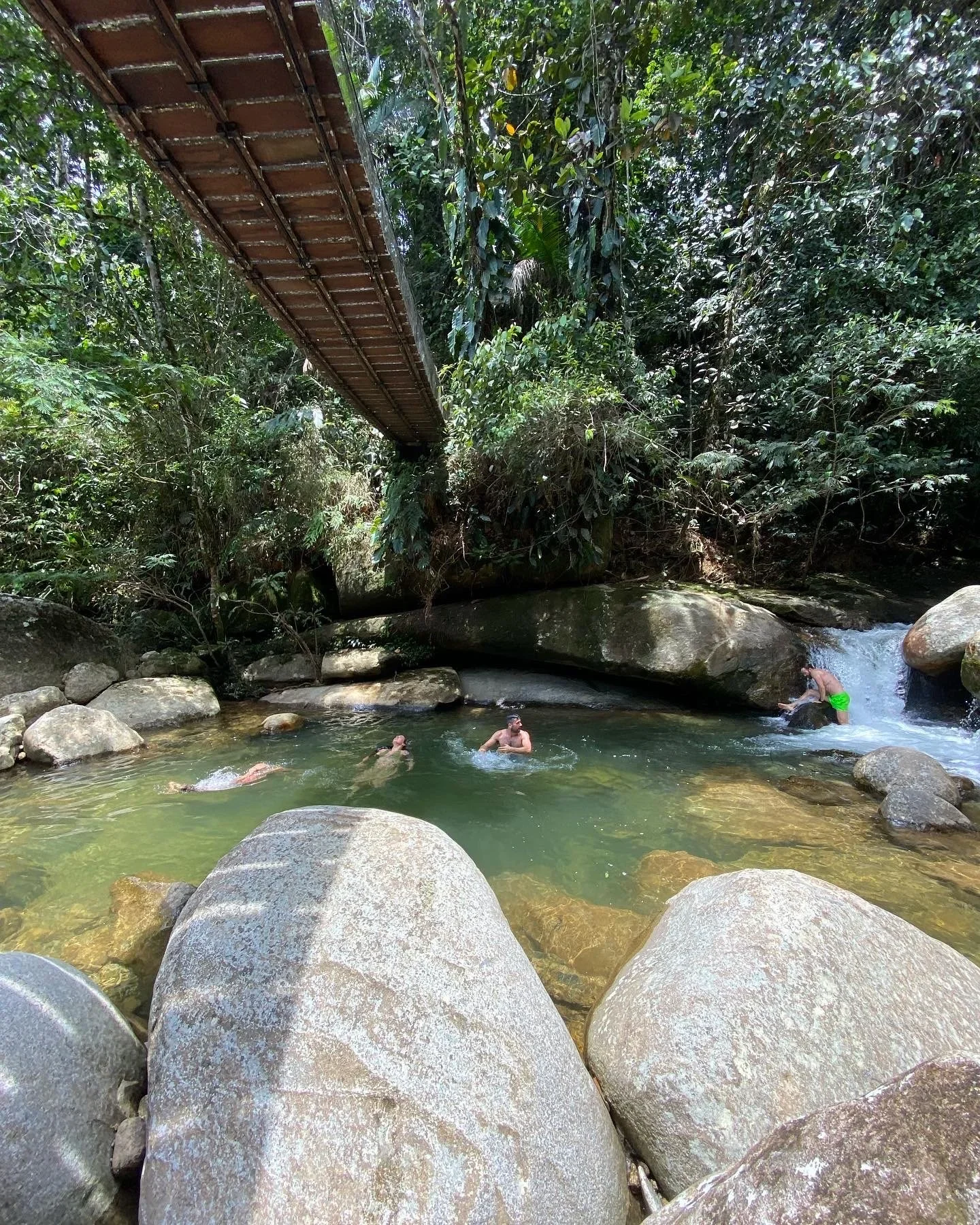 People swimming and relaxing in a river beneath a bridge in a lush jungle setting, with large rocks in the water.