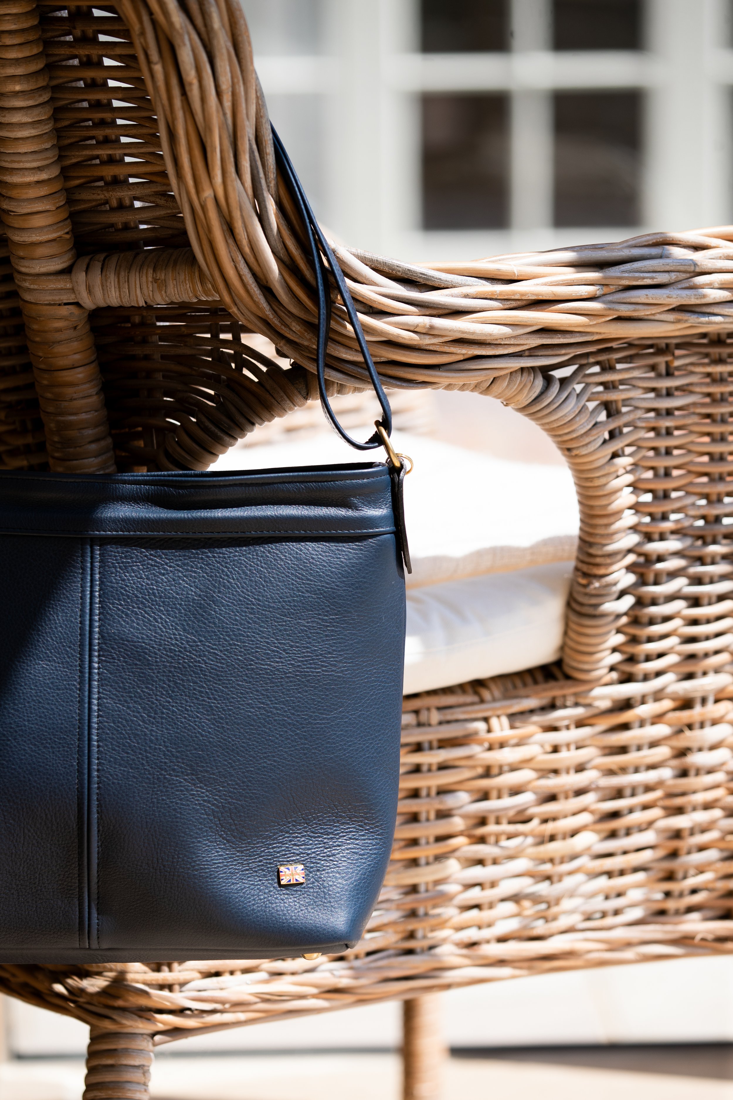 A navy leather Kate Negus handbag hanging from the armrest of a woven wicker chair outdoors.