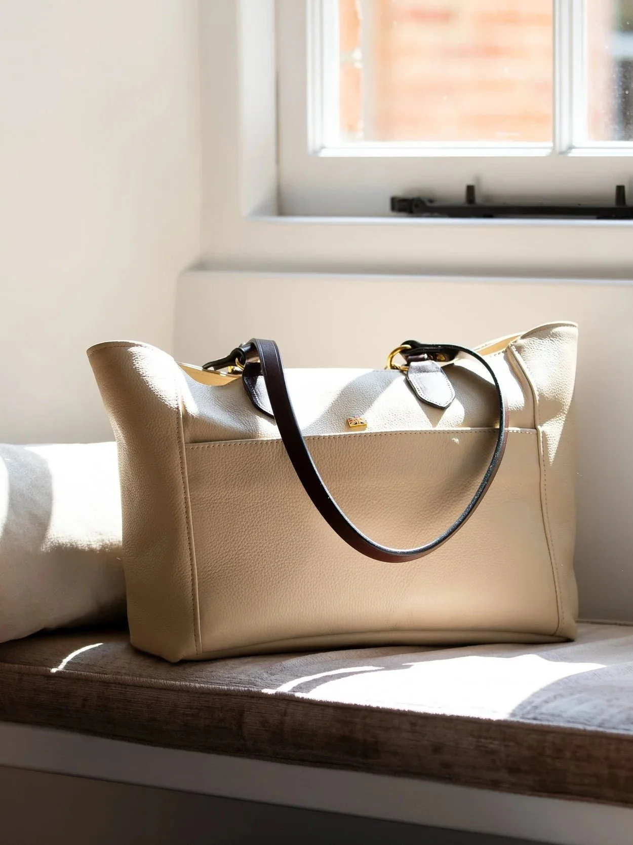 A beige leather Kate Negus tote bag with black straps sitting on a windowsill next to a white cushion, sunlight shining through the window.