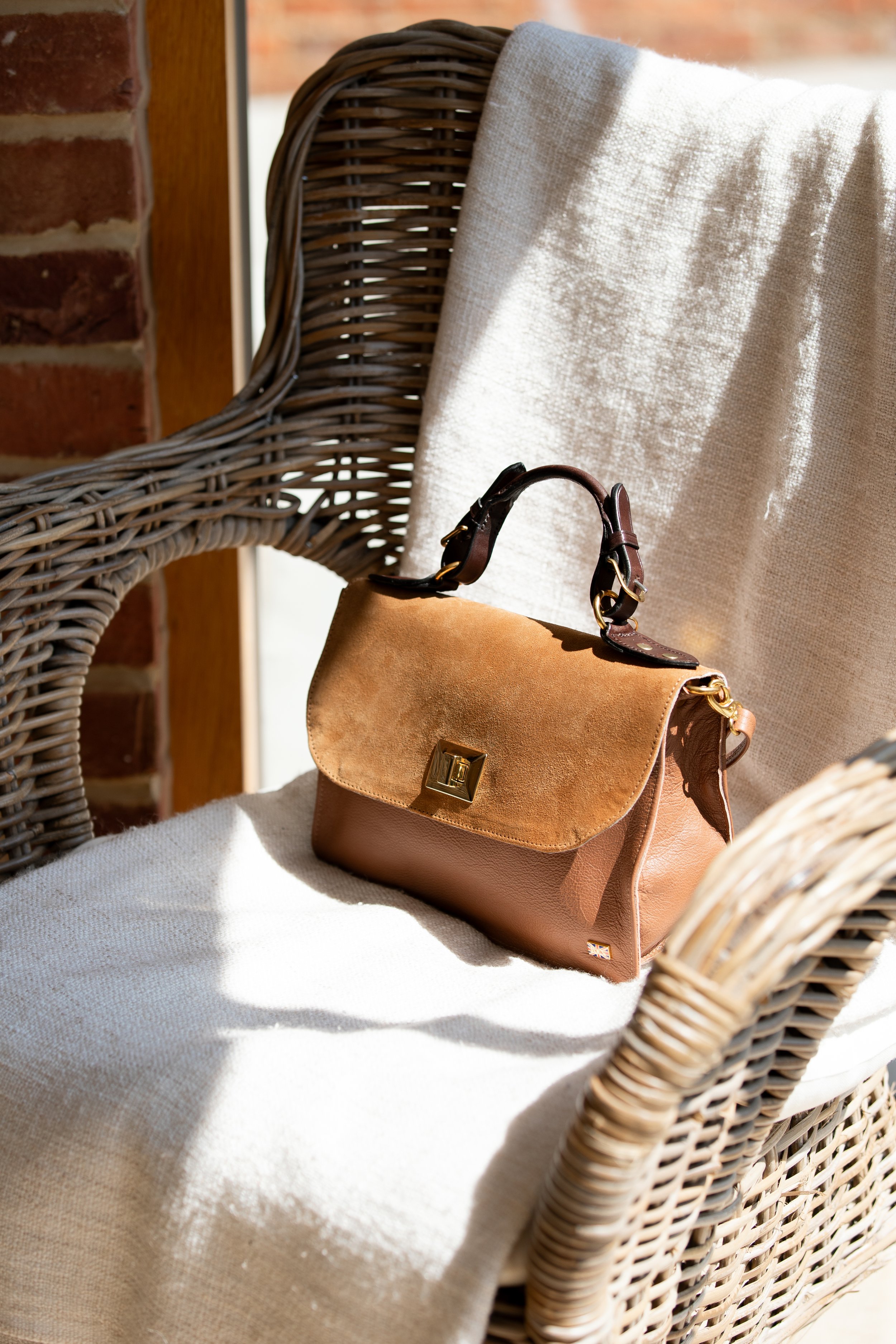 A tan leather Kate Negus handbag with a gold clasp resting on a white cushion on a wicker chair, near a brick wall and window.