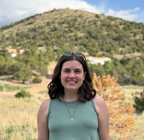 Smiling woman with dark hair and sunglasses on her head standing outdoors in a scenic landscape with mountains and trees.