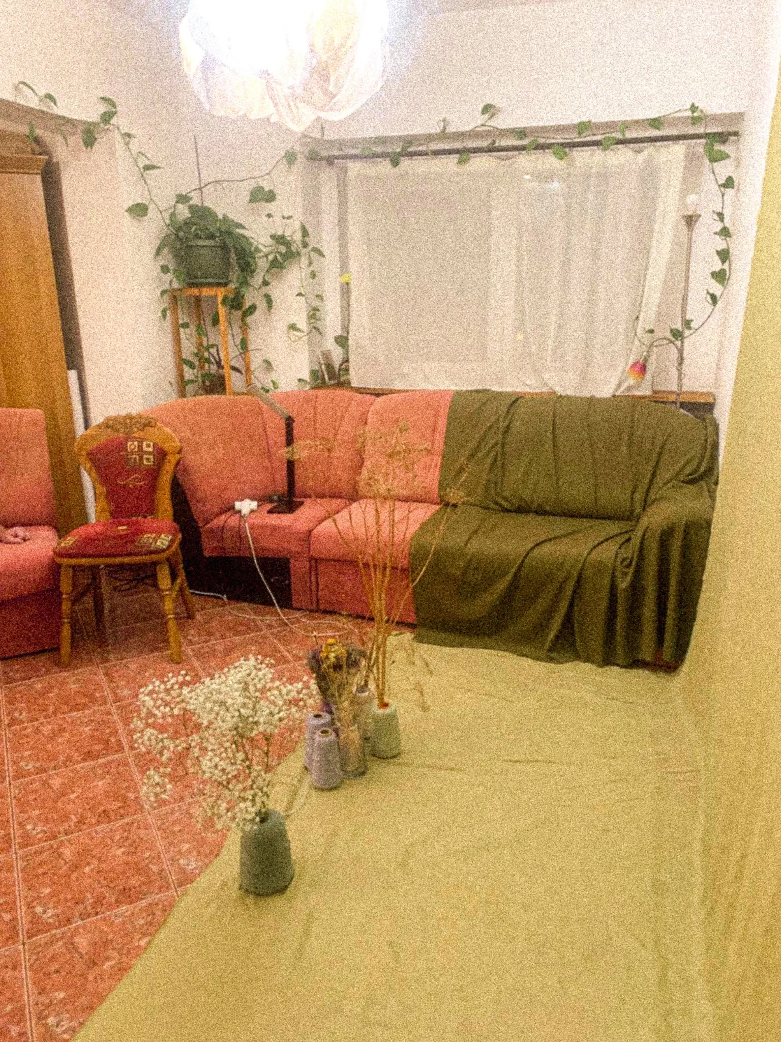 A living room with a pink and green sectional sofa, a small wooden table, and various plants, including a large plant on a stand and a potted plant hanging with vines. The floor has red tiles and the wall has a window covered with a white curtain. There is a ceiling lamp with a paper lampshade.