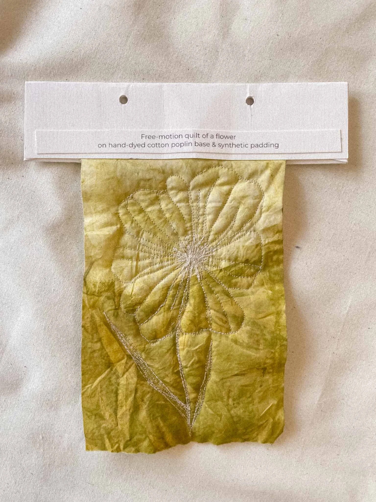 A piece of gold-colored fabric with an embroidered flower and a white label at the top that reads, "Free-motion quilt of a flower on hand-dyed cotton poplin base & synthetic padding."