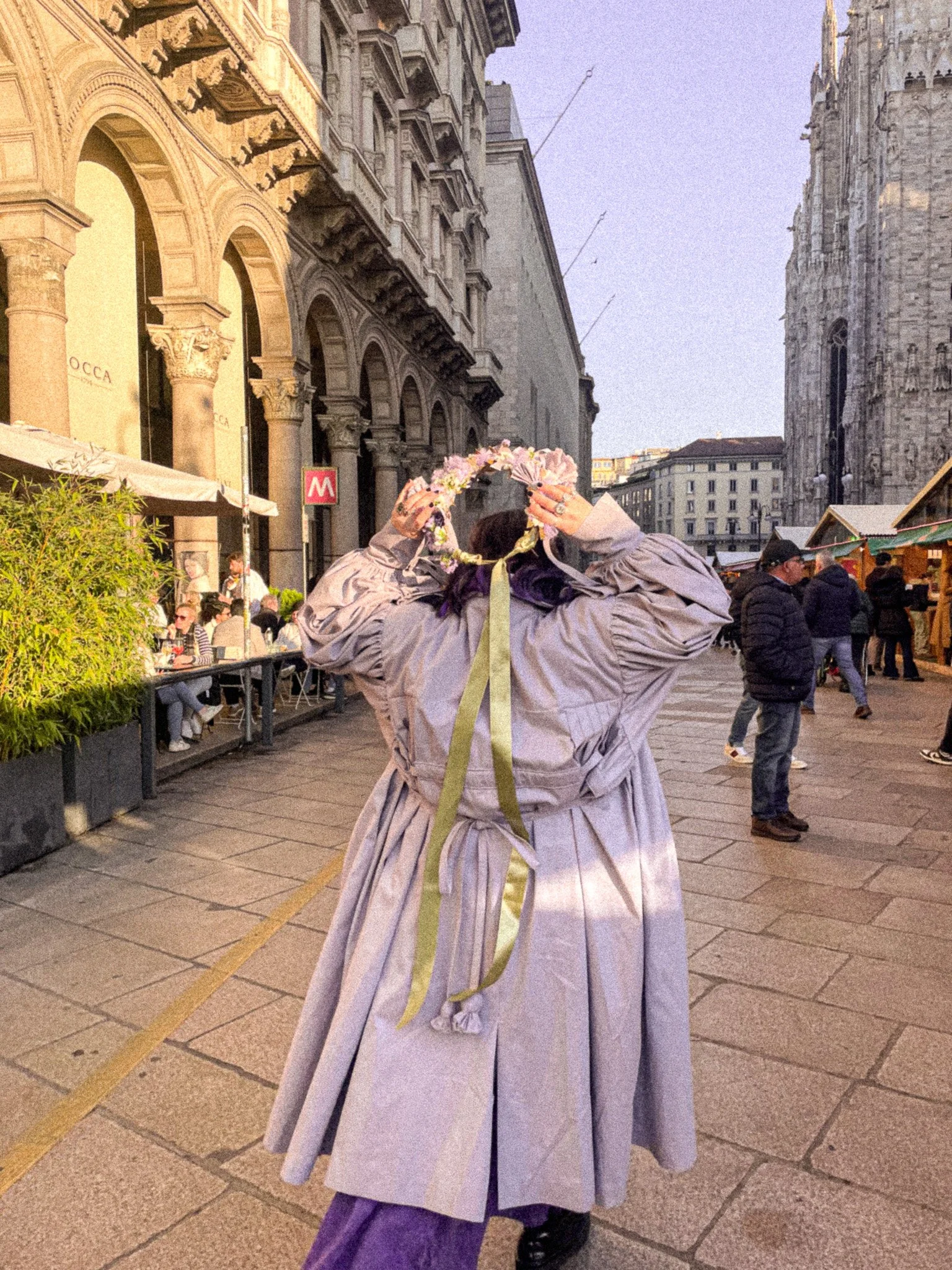 Person wearing a long gray dress and purple pants, adjusting a floral crown on their head in a city street with historic buildings and onlookers.