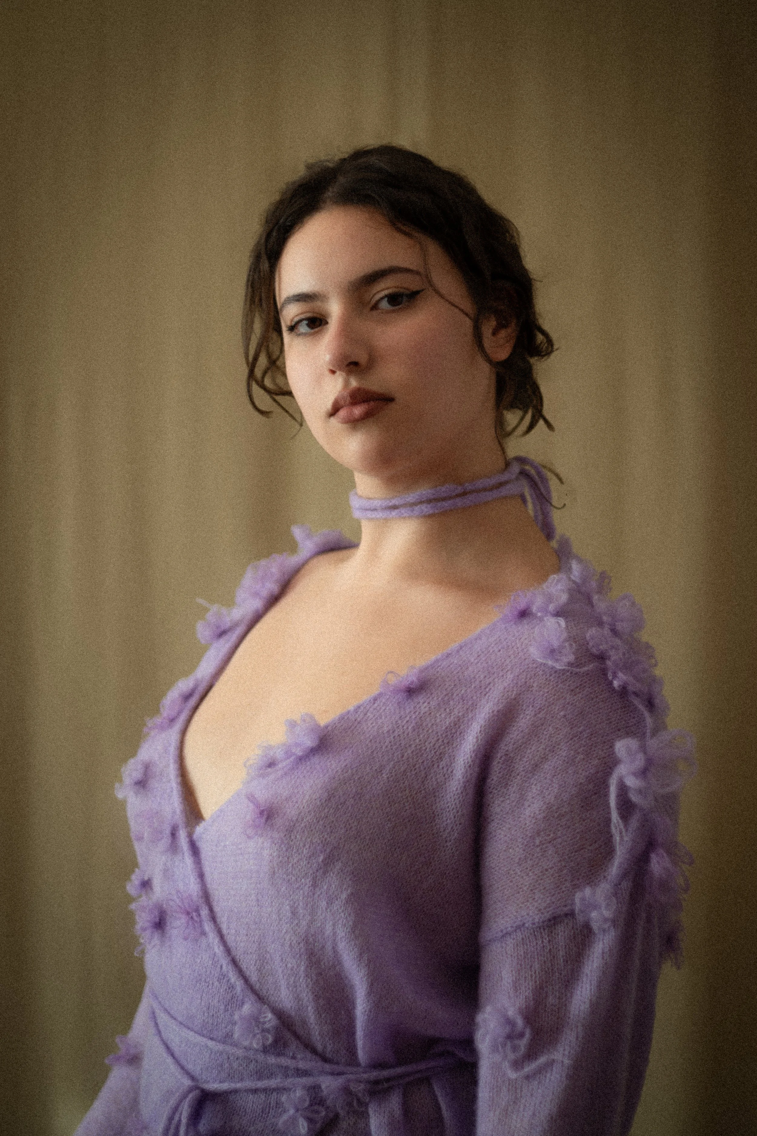 A woman with short dark curly hair wearing a lavender sweater with ruffled details and a matching ribbon choker, posing against a neutral background.