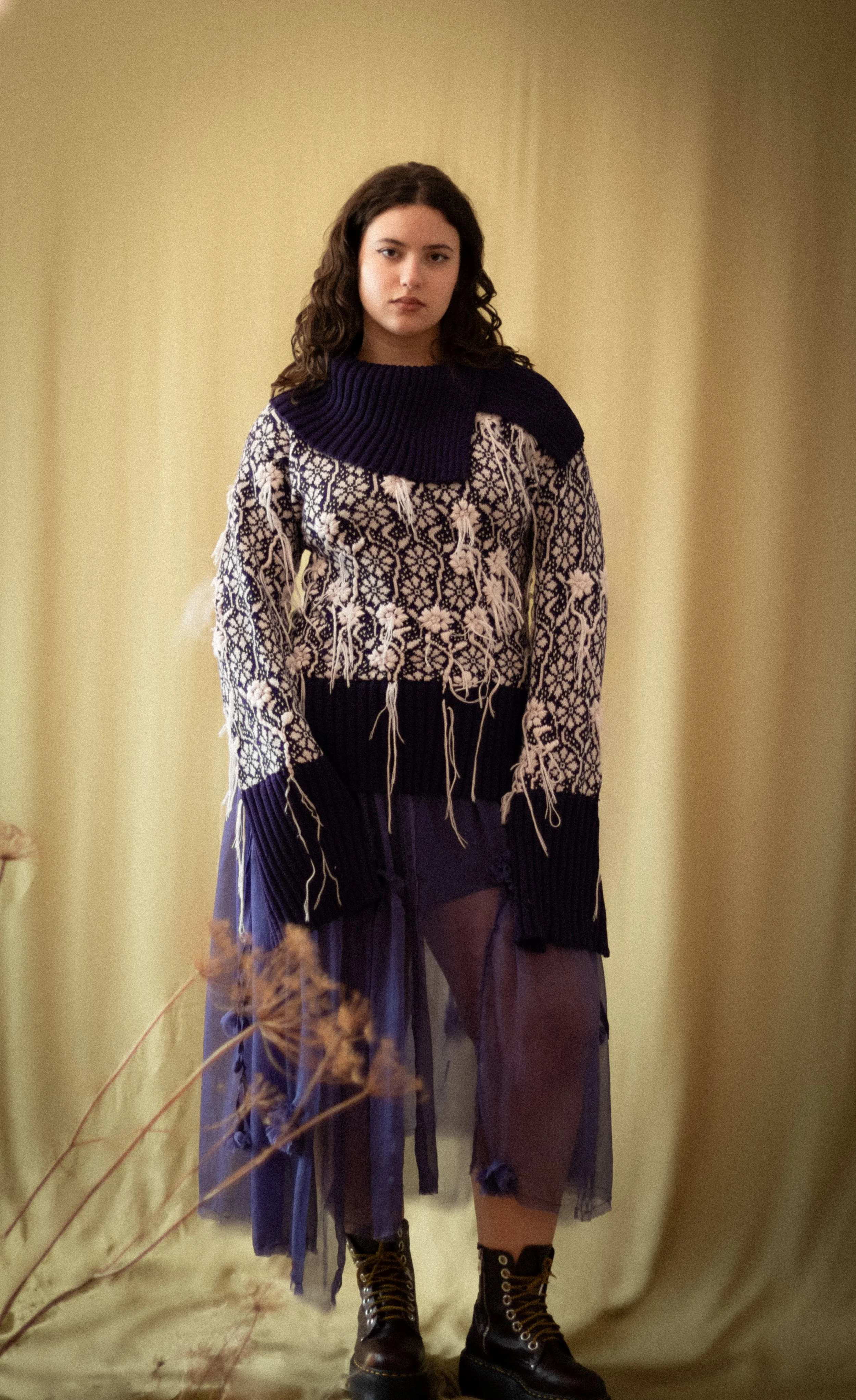A young woman with curly dark hair, wearing a layered outfit including a patterned top with raw edges, a purple skirt, and lace-up boots, standing in front of a plain background with dried plants in the foreground.