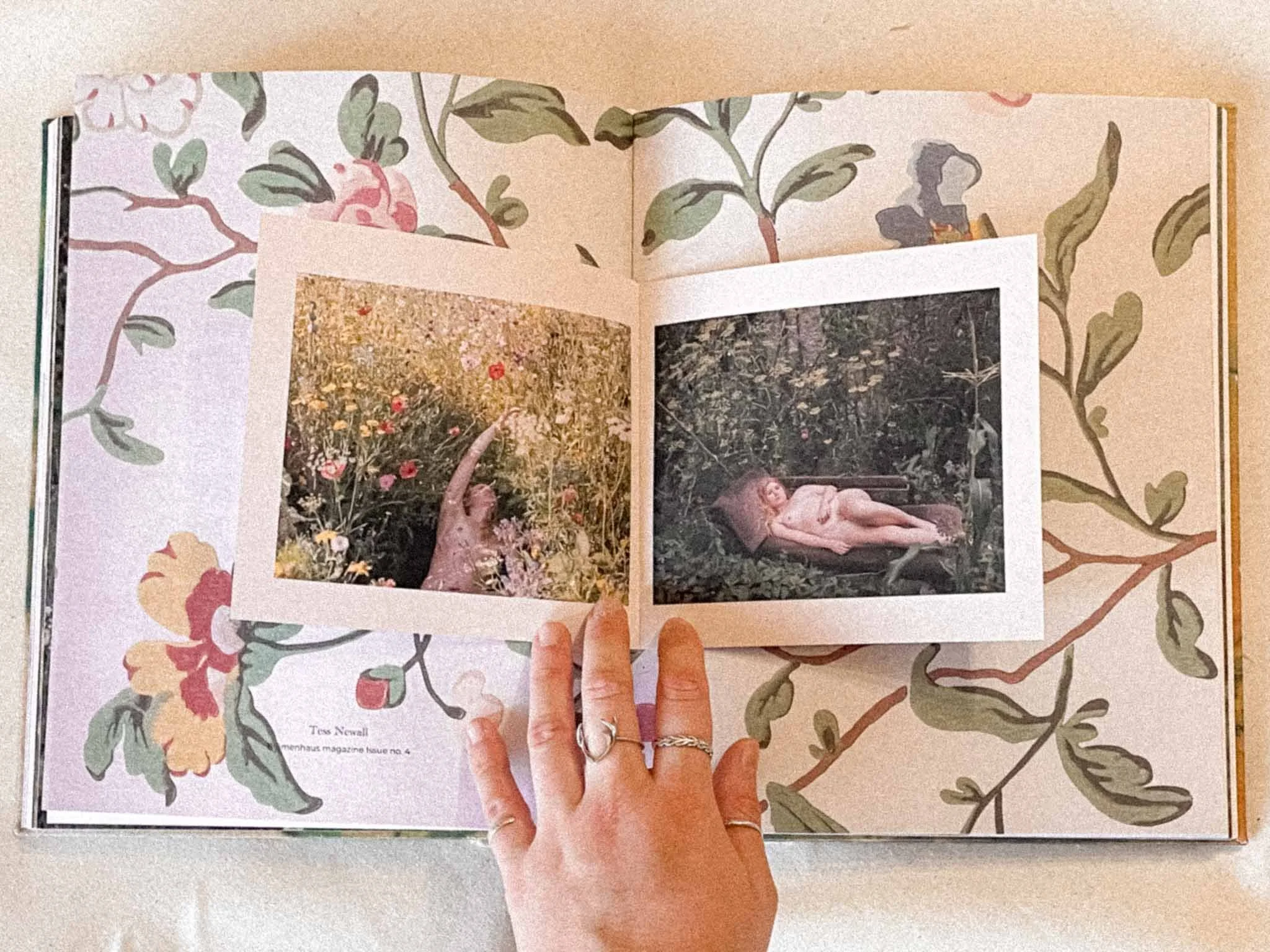 Open magazine with floral border and two photographs, one of a person lying on the floor with a background of trees and flowers, and the other of a woman sitting among flowers.