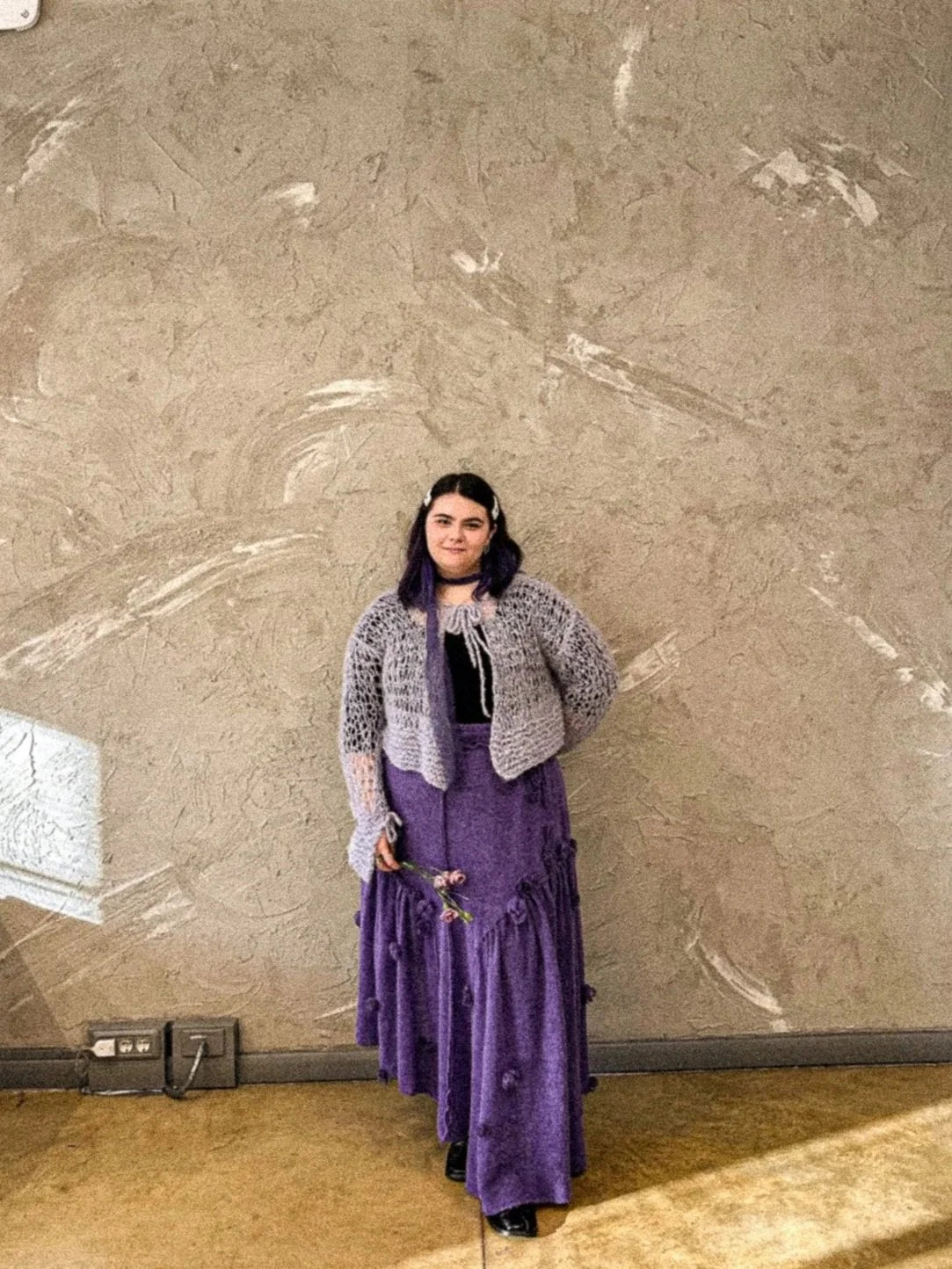 A woman with dark hair and purple highlights, dressed in a purple skirt and a gray, open-knit jacket, standing against a beige textured wall, holding a small bunch of flowers.