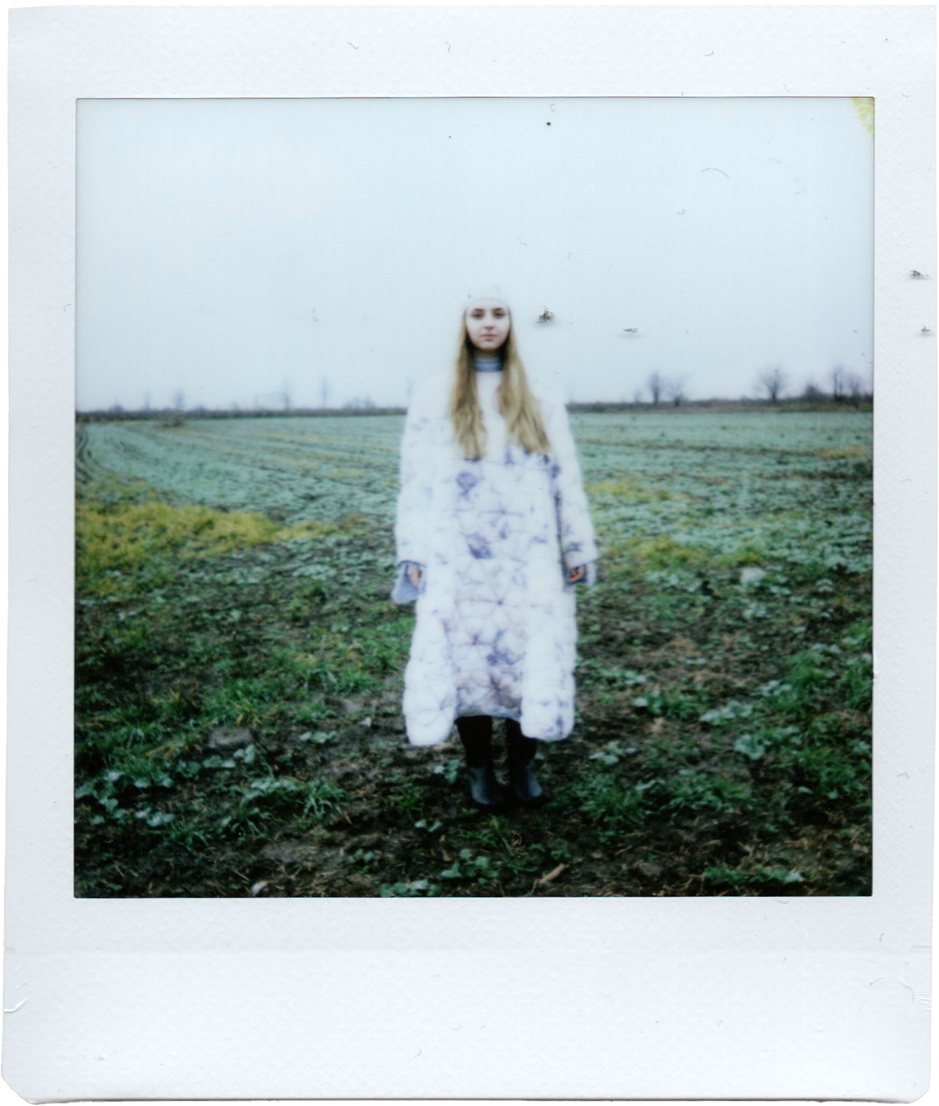 A young woman with long blonde hair stands outdoors in a grassy field, wearing a long, white, tie-dye coat and black boots, with trees in the background under a cloudy sky.