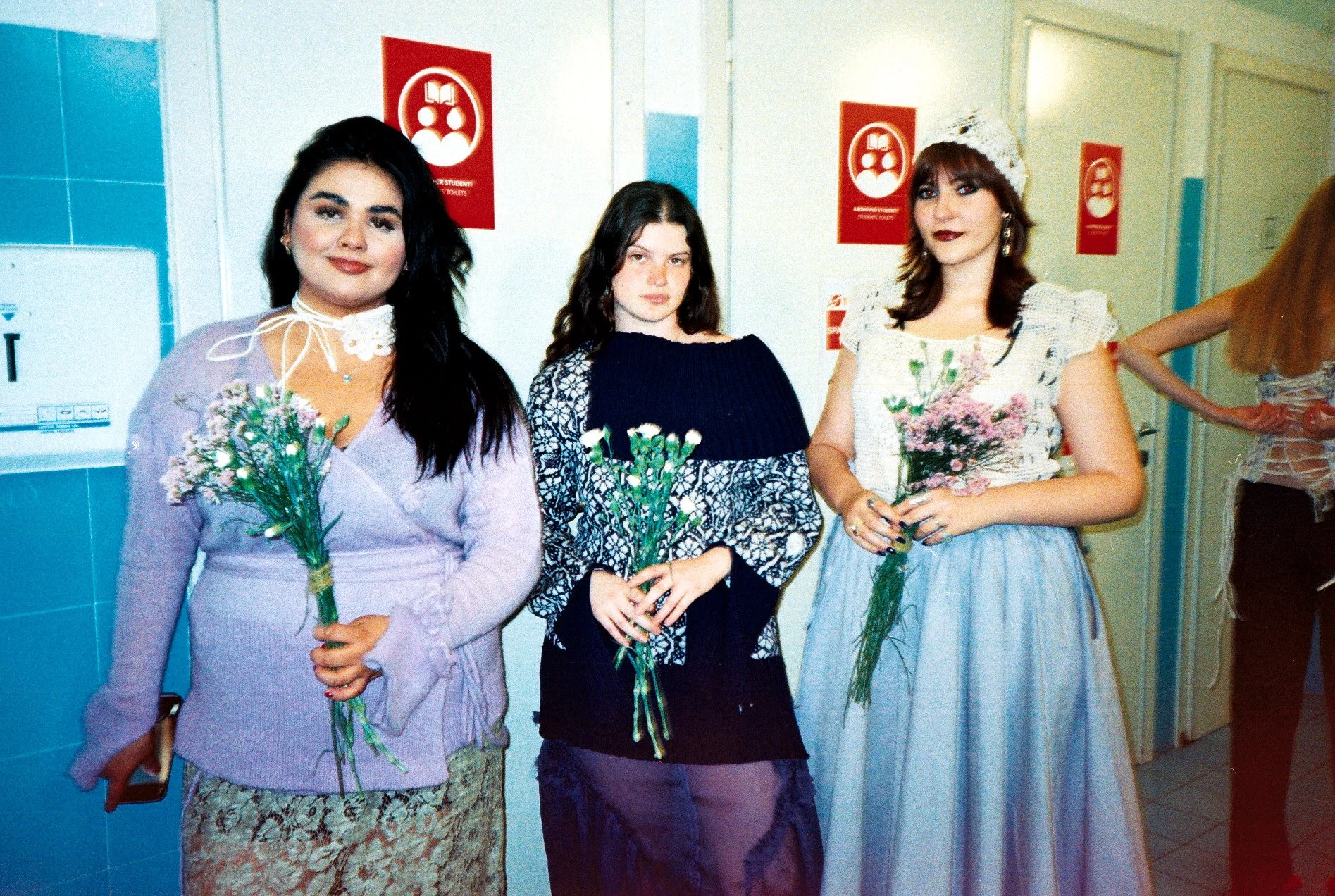 Three young women standing in a hallway, each holding a bouquet of flowers, with a background of blue and white tiled walls and red signs indicating restrooms.