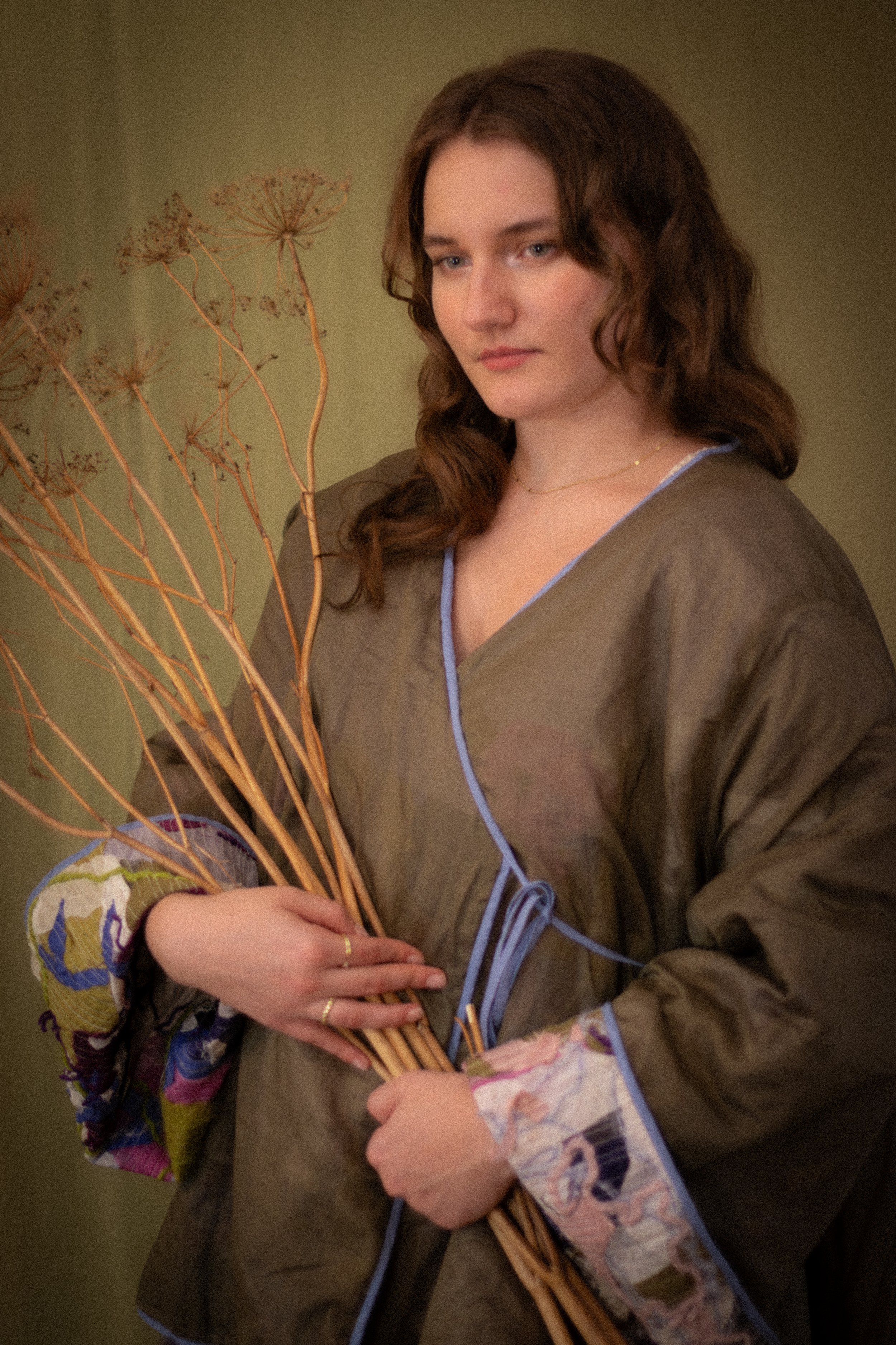 A woman with long, wavy brown hair holds a bunch of dried plant stems and a colorful round object, wearing a loose brown top with blue piping, standing against a greenish background.