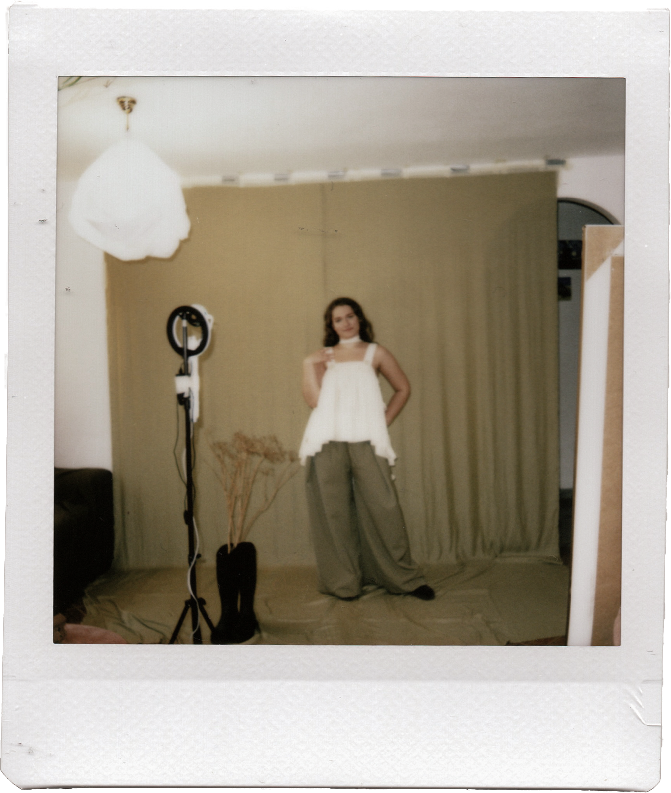 A woman standing in front of a green backdrop in a photography studio, wearing a white top and beige pants, with a professional studio light to her left and a plant nearby.