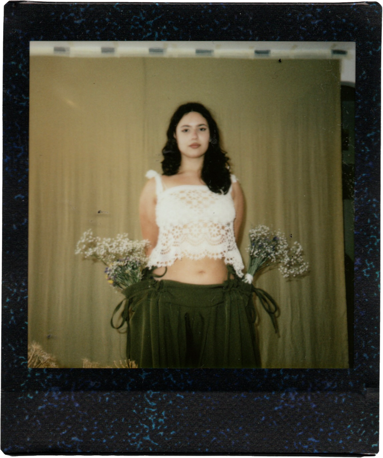A young woman with dark, wavy hair, wearing a white crochet crop top and green pants, standing in front of a beige curtain, holding bunches of small white flowers in her pockets.
