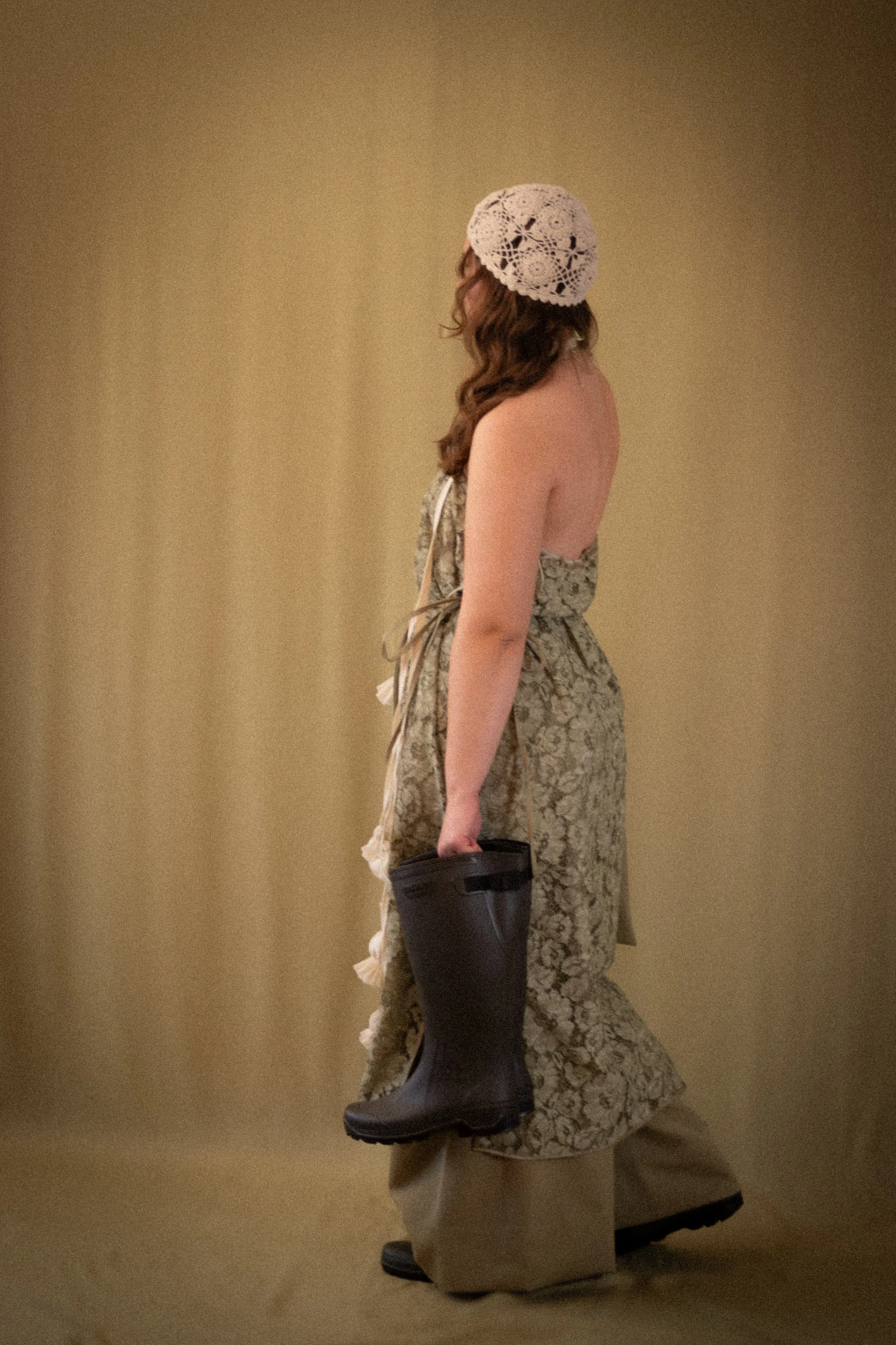 A woman dressed in a patterned strapless dress, wearing a white crochet hat, black rubber boots, and carrying a black blog in her right hand, standing against a plain beige background.